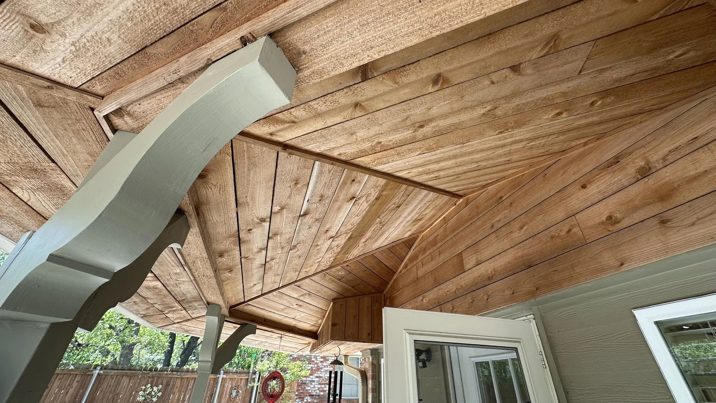 Wood-paneled ceiling and part of a windowed door on an outdoor porch with visible wooden fence and trees in the background.