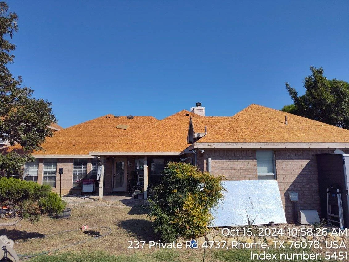 A house with a brown roof and brick walls. There is a tree on the left and a bush in front of the house. It is a sunny day with a clear blue sky. Construction or renovation materials are visible in the yard.