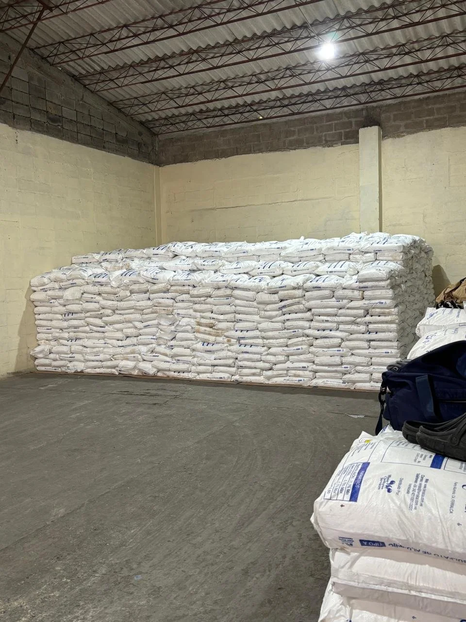 A stack of white bags filled with a material stored in a room with unfinished concrete walls and a metal roof. There are also a few more bags and backpacks on the floor to the right.