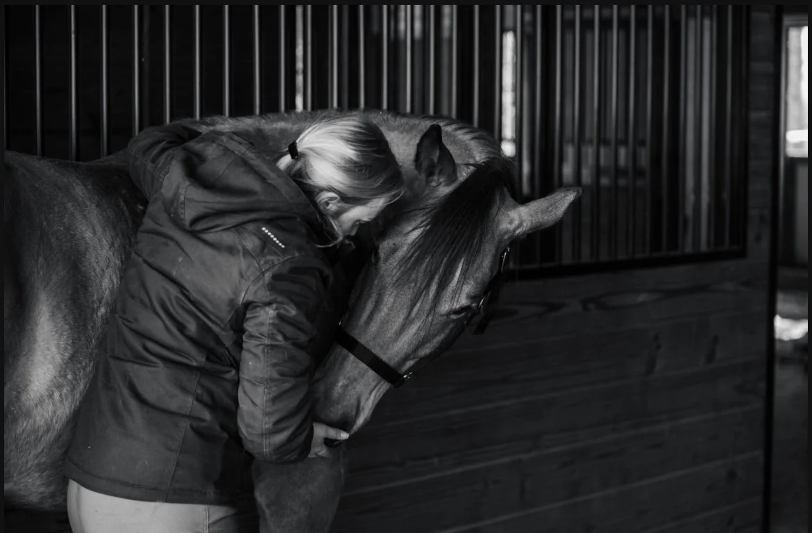 A woman hugging and pressing her face against a horse's head inside a wooden stable.