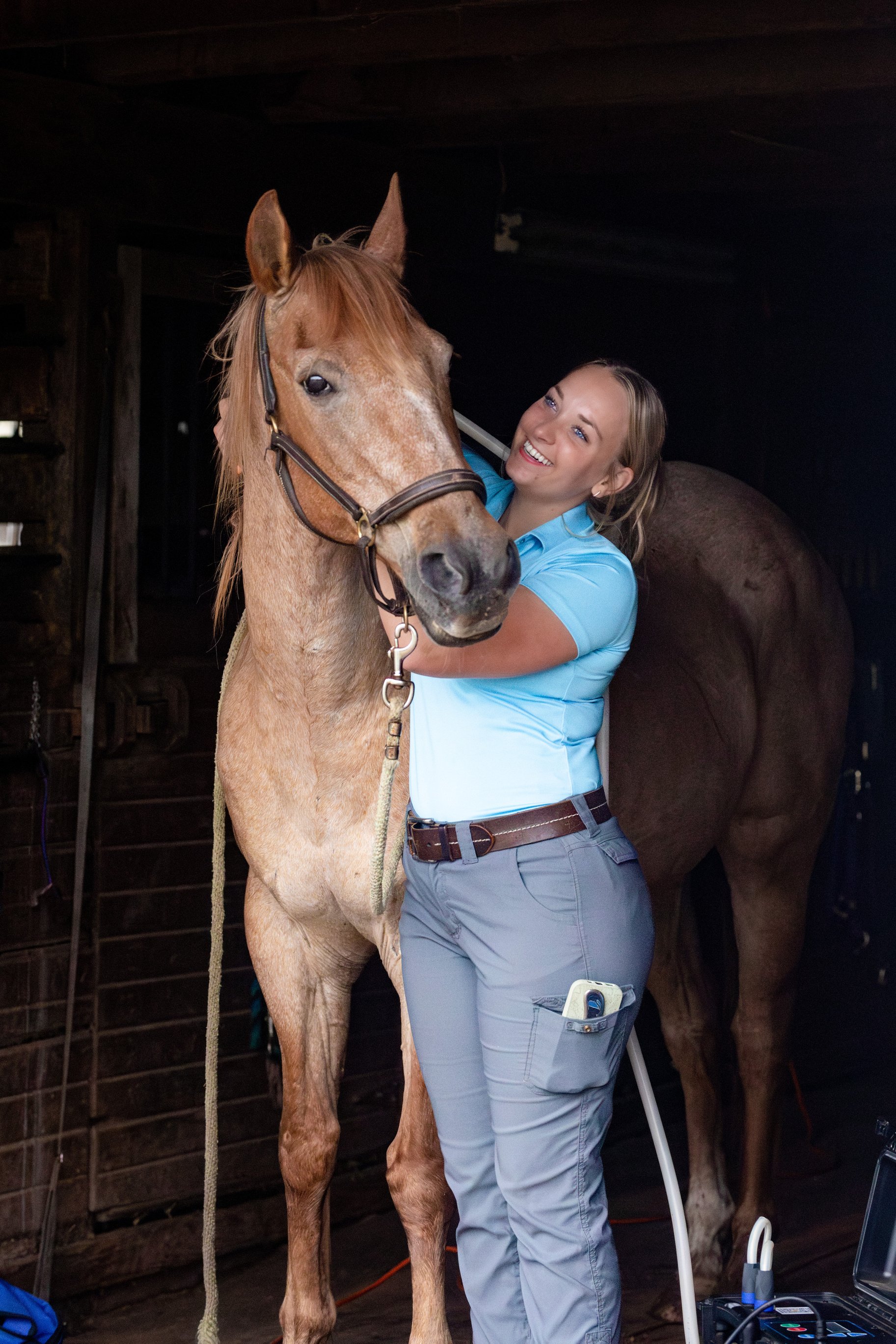 A woman smiling and hugging a light brown horse inside a barn.
