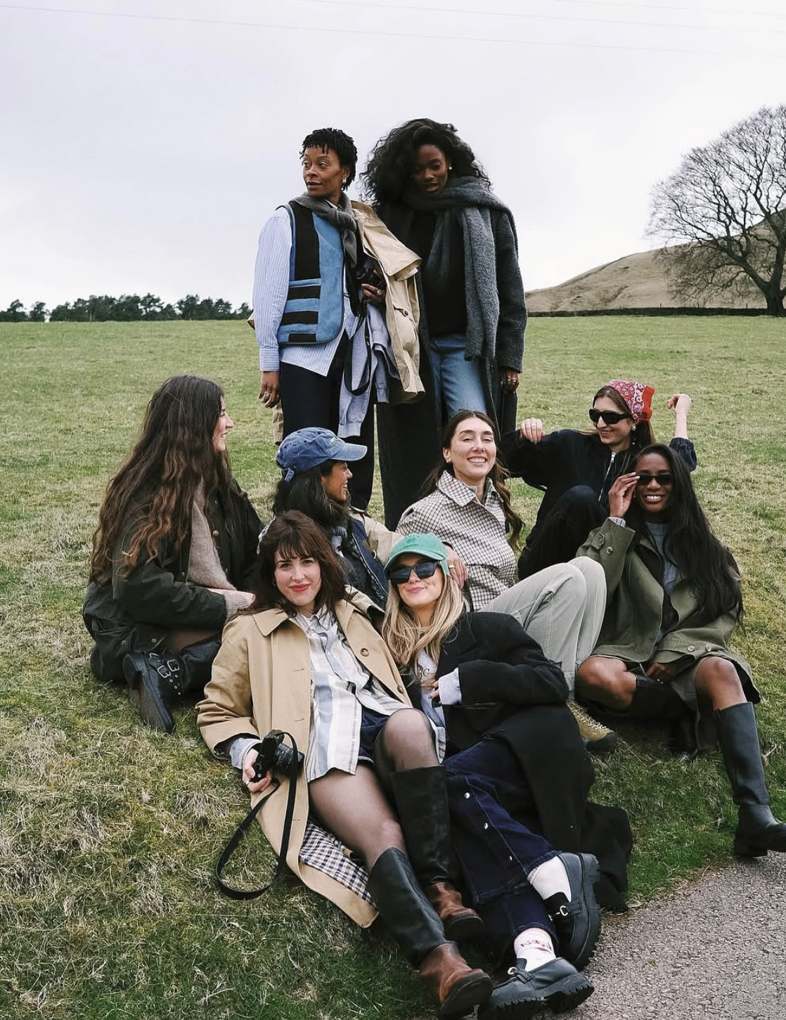 Group of women outdoors on a grassy hill, some sitting and some standing, smiling and posing for a photo, with a cloudy sky and a tree in the background.
