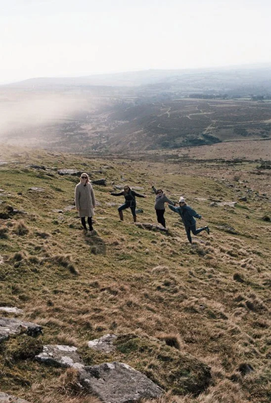 A dining table set for a meal with plates, napkins, and glasses of white wine or champagne. The table has a large floral centerpiece with white flowers and green foliage. There are candles and modern black and wooden decorative objects on the table. The background features a rustic stone fireplace with a brick mantel.