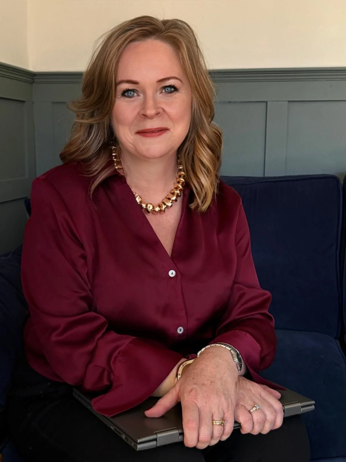 Helen Grant of Diolta Estate Agenc wearing a maroon satin blouse,  a gold bracelet, and a wristwatch. She has rings on her fingers and is holding a closed laptop on her lap. The background features panelled walls in muted green and beige colours.