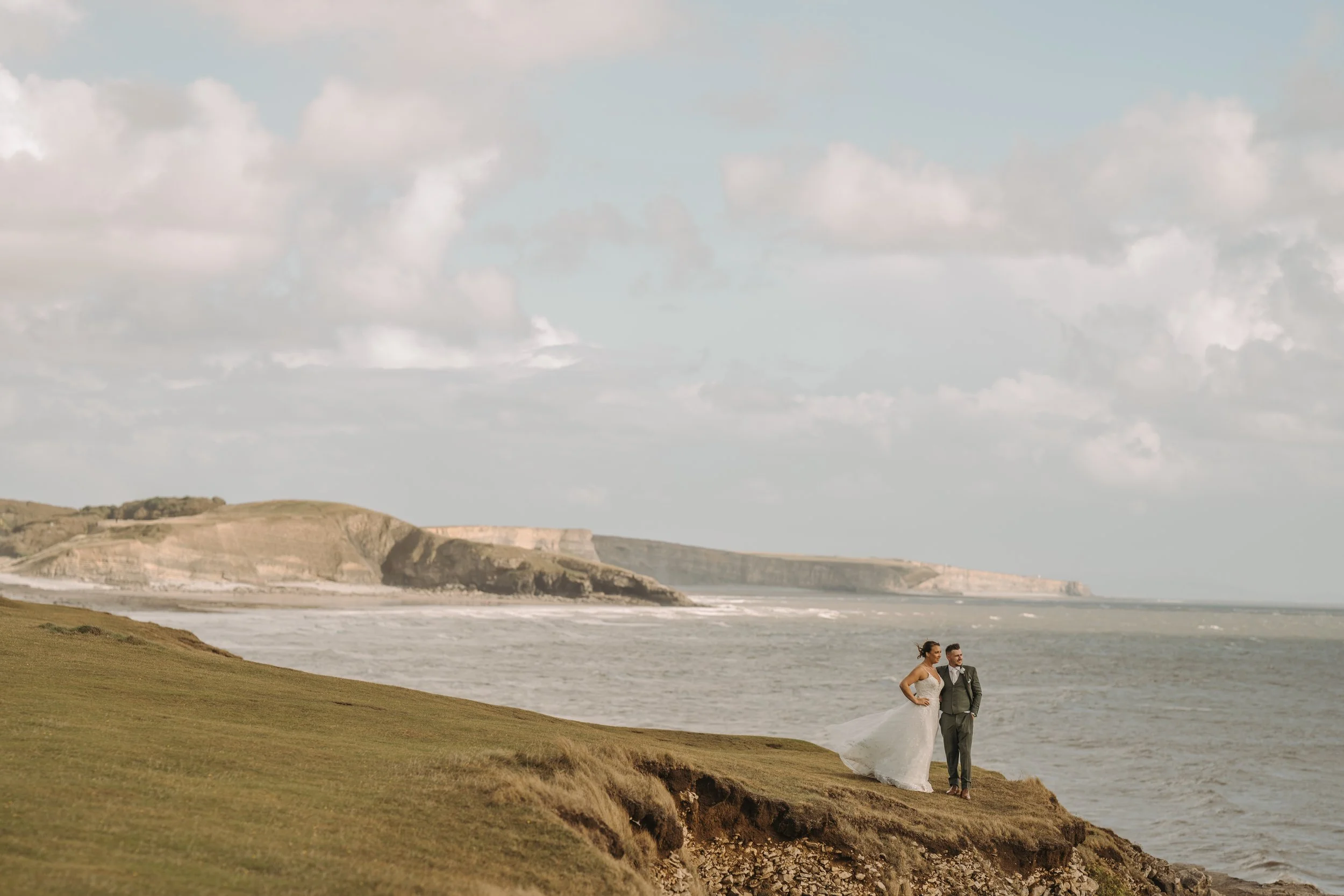 Bride and groom standing on grassy cliff overlooking the ocean with distant cliffs and cloudy sky.