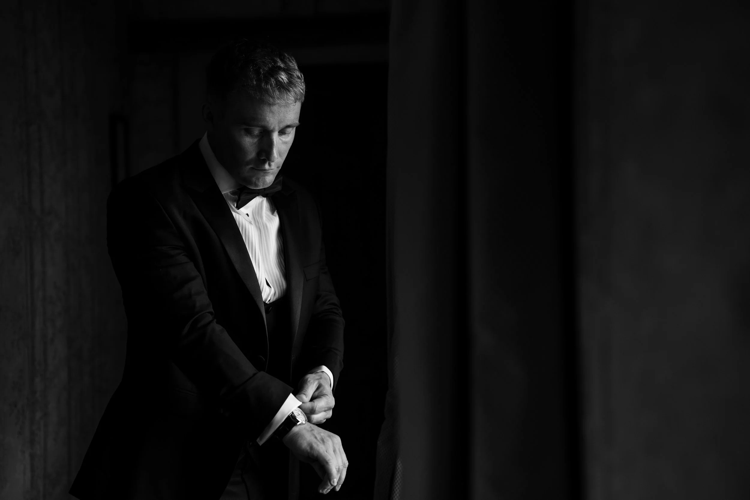 A man dressed in a tuxedo with a bow tie adjusting his sleeve in a dimly lit room.