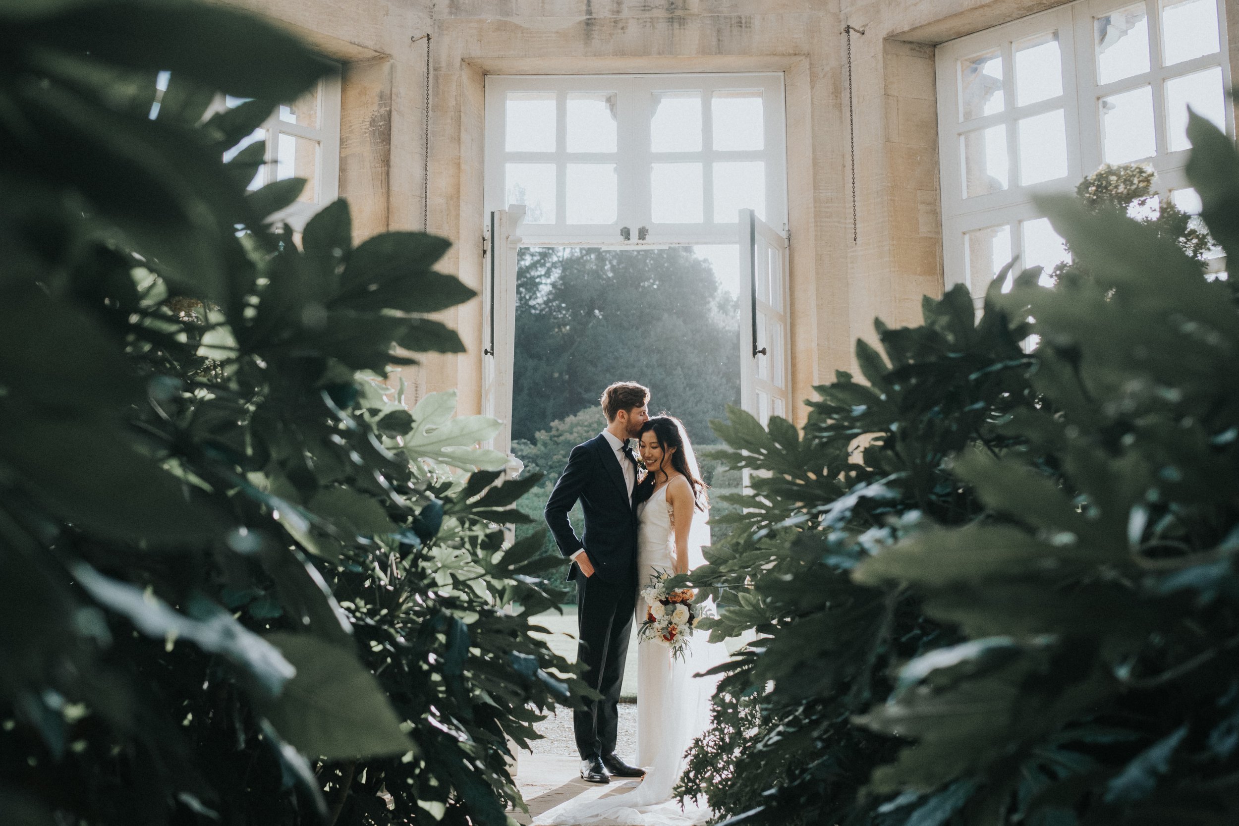 A bride and groom standing close together inside a building with large open windows, framed by green foliage in the foreground, during daylight.