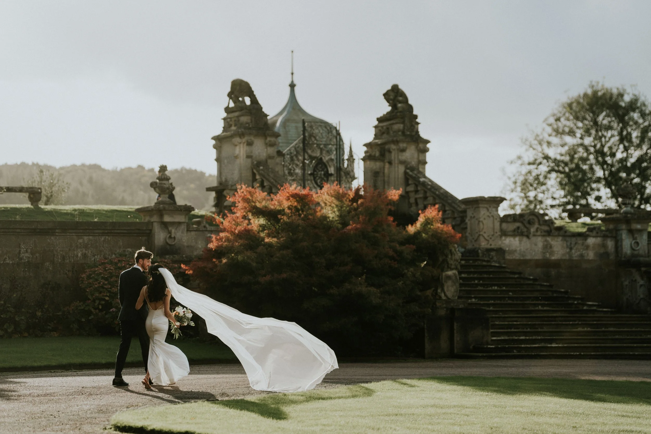 Bride and groom walking together outdoors near historic stone building with stairs and trees, sunset lighting.