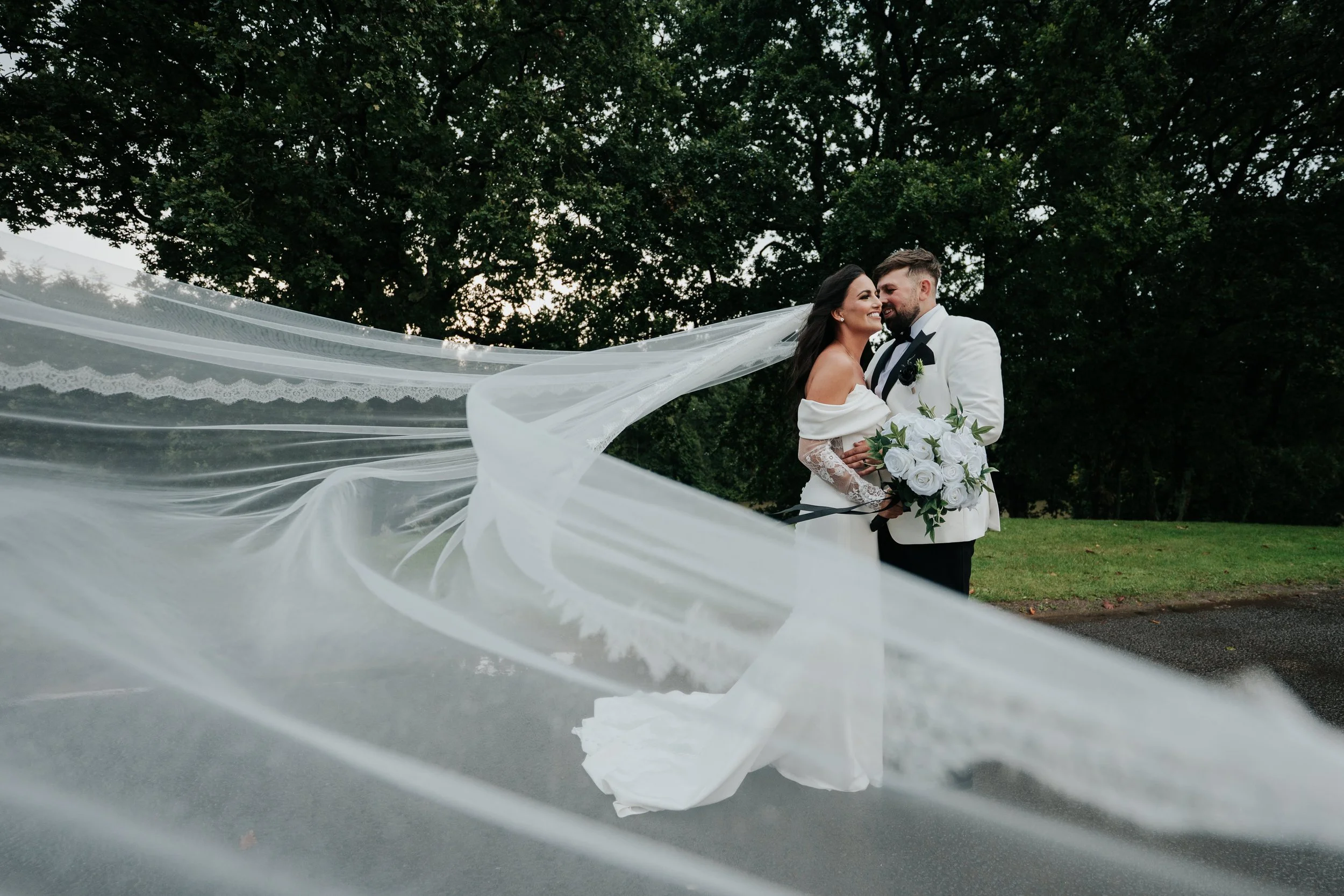 A bride and groom happily embrace outdoors, with the bride's flowing veil swirling around them and trees in the background.