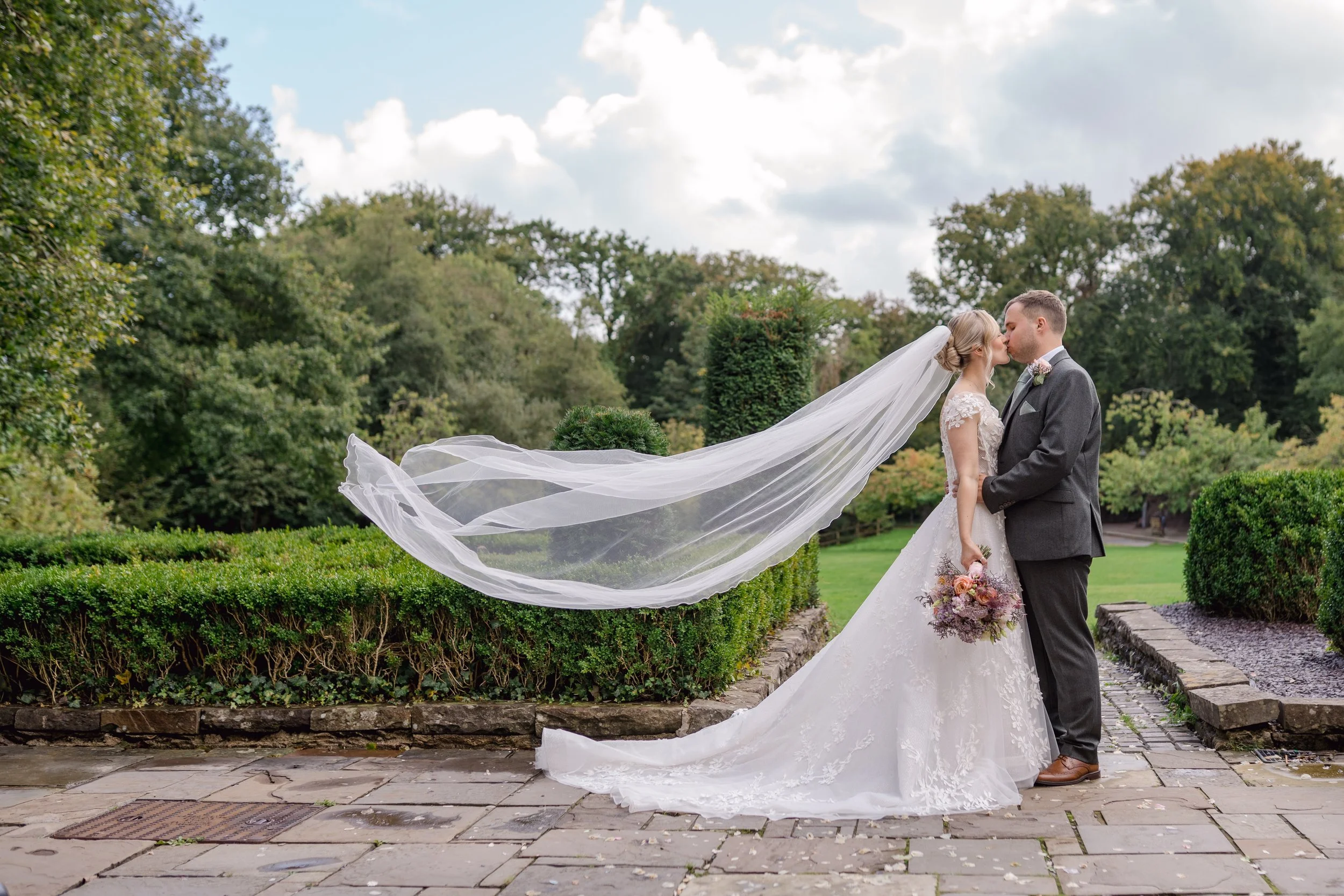 A bride and groom sharing a kiss outdoors, with the bride holding a bouquet and her veil flowing in the breeze against a background of green trees and cloudy sky.