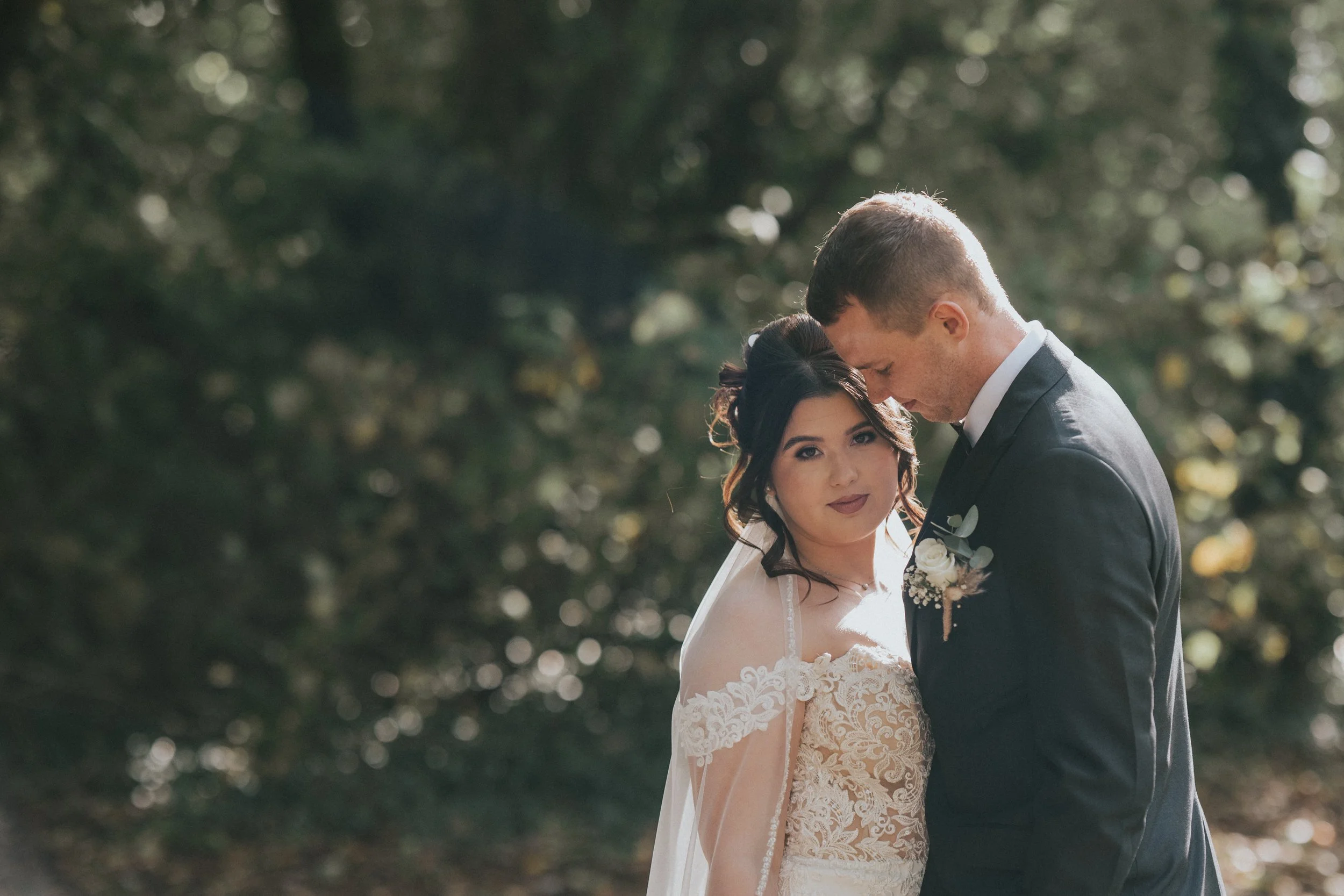 A bride and groom standing close together outdoors, with the groom gently resting his forehead on the bride's hair, surrounded by greenery and sunlight filtering through trees.