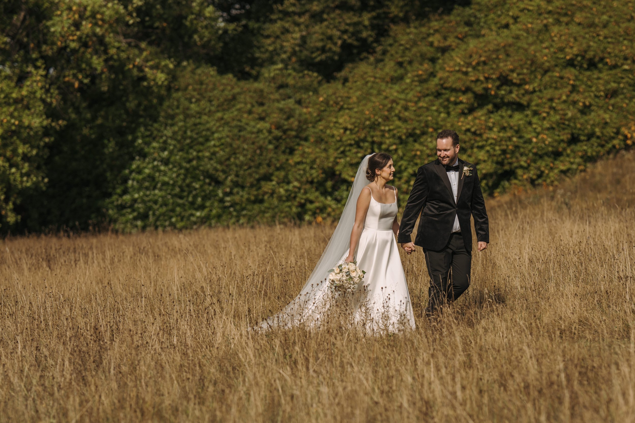 A bride and groom walking hand in hand through a grassy field, with trees in the background, on their wedding day.