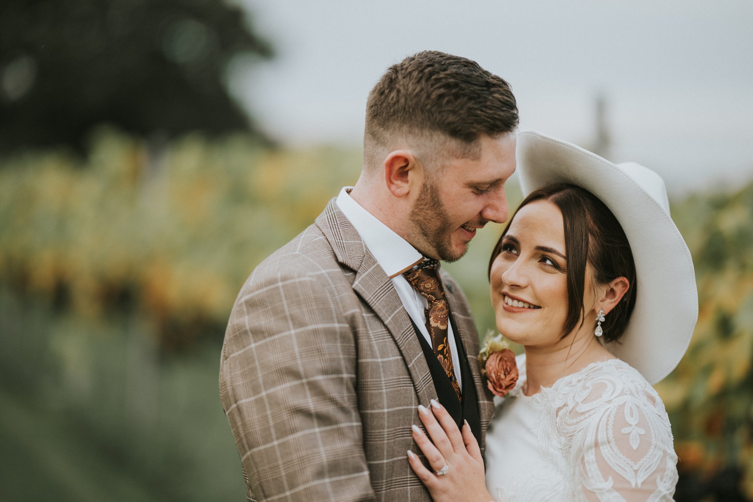 A couple on their wedding day, with the groom in a checkered suit and the bride in a white dress and wide-brimmed hat, smiling at each other outdoors.