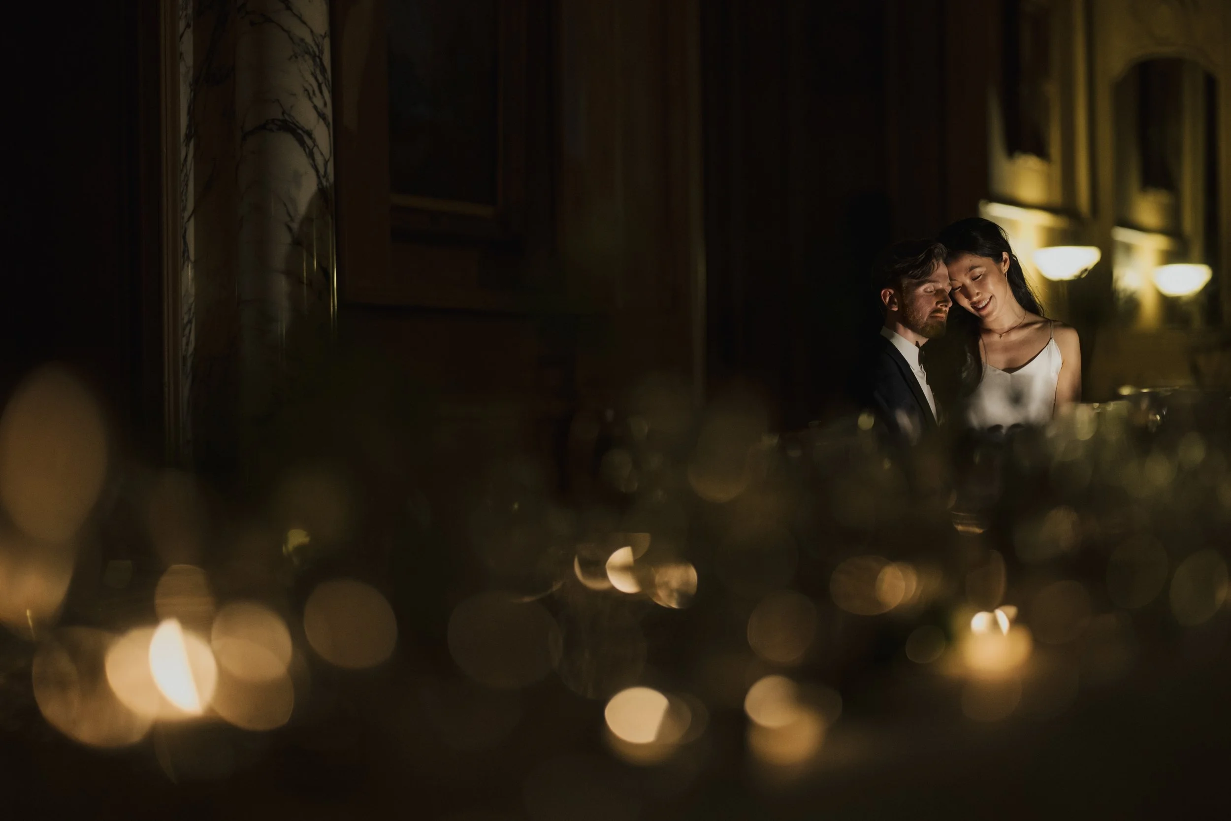 A couple at a dimly lit dinner table, with warm yellow lighting and blurred foreground lights, sharing an intimate moment.