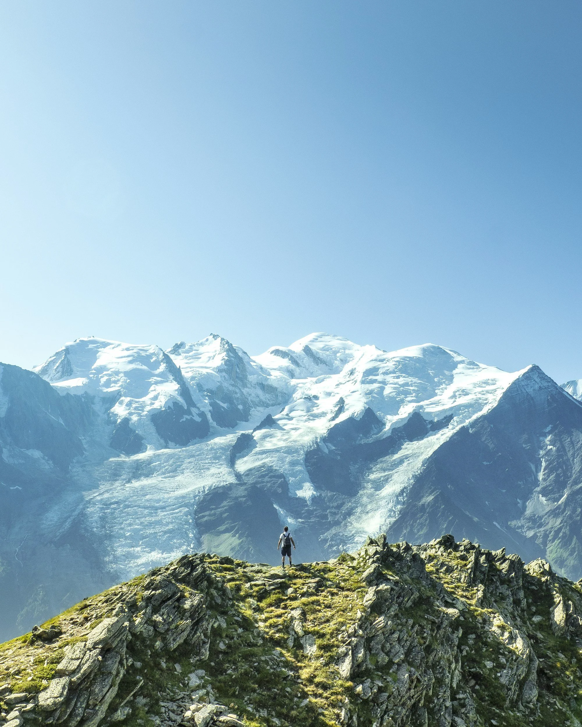 A person standing on a rocky hilltop overlooking snow-capped mountains and glaciers under a clear blue sky.