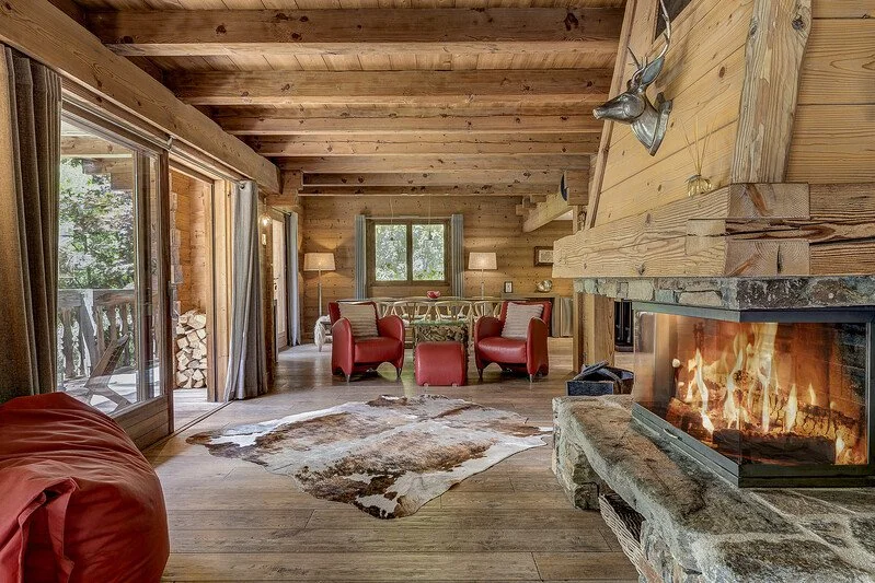 Cozy chalet living room with wood-paneled walls and ceiling, a roaring fire in a stone fireplace, two red armchairs, a cowhide rug, large sliding glass doors leading to an outdoor balcony, and a window behind the furniture.
