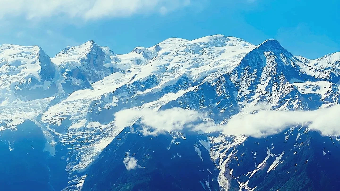 Snow-covered mountain peaks with clouds around the slopes under a blue sky.