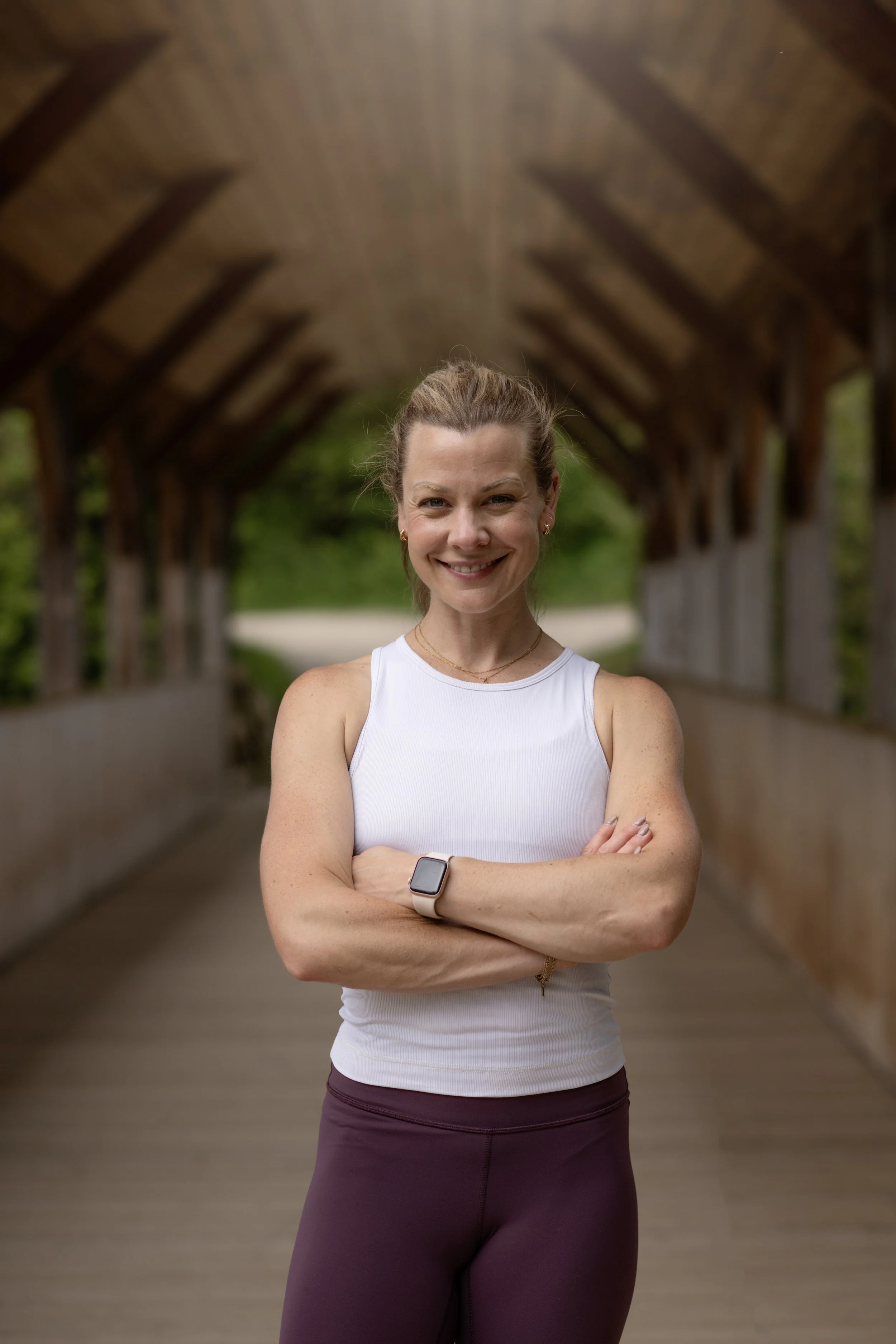 A smiling woman in athletic wear standing in a covered outdoor pathway with arms crossed.