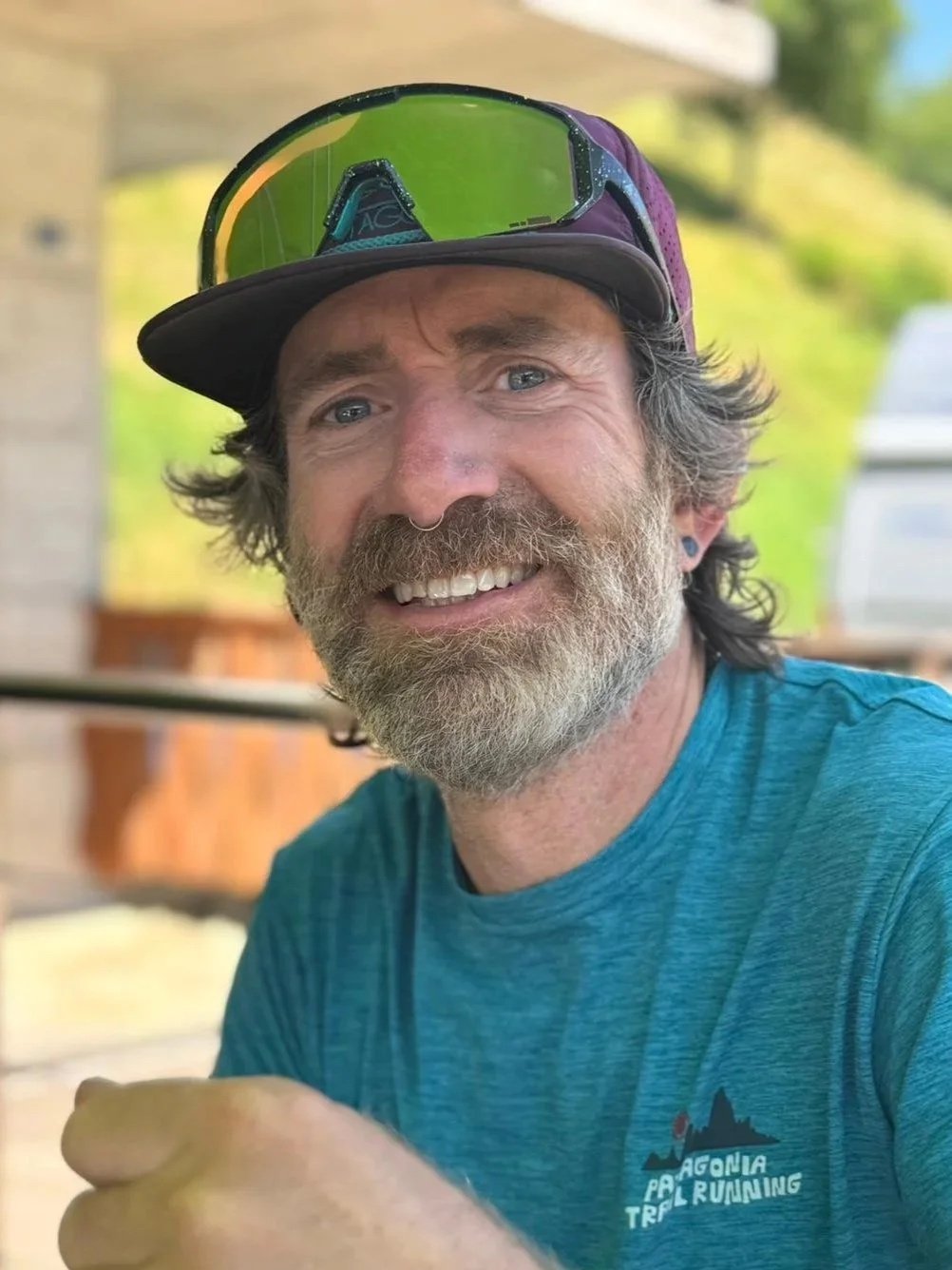 A smiling man with a beard and mustache, wearing a blue shirt, a cap, and sunglasses on his forehead.