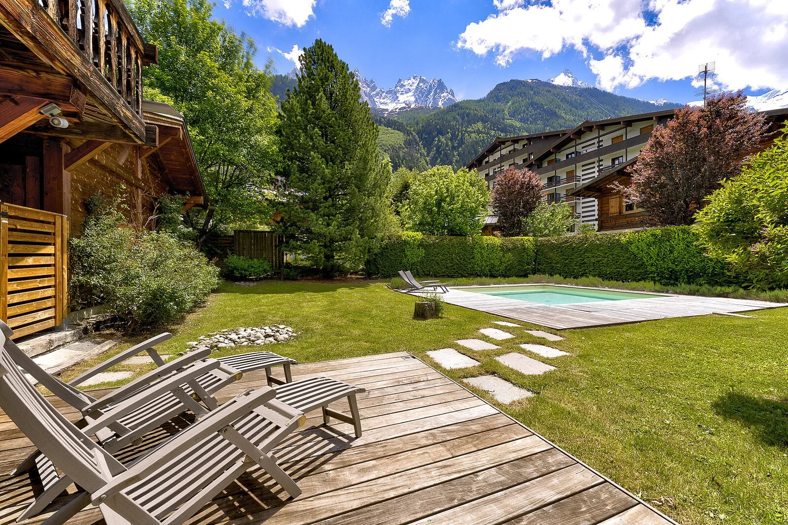 A backyard with a pool, wooden decking, lounge chairs, lush green grass, trees, hedges, and a mountain backdrop with snow-capped peaks.