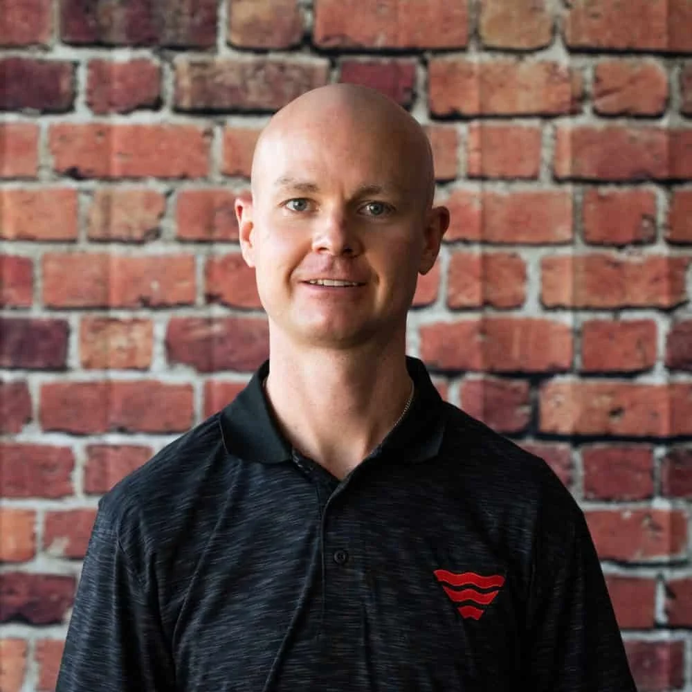A man with a shaved head and light skin, wearing a black shirt with a red wave logo, standing in front of a brick wall.