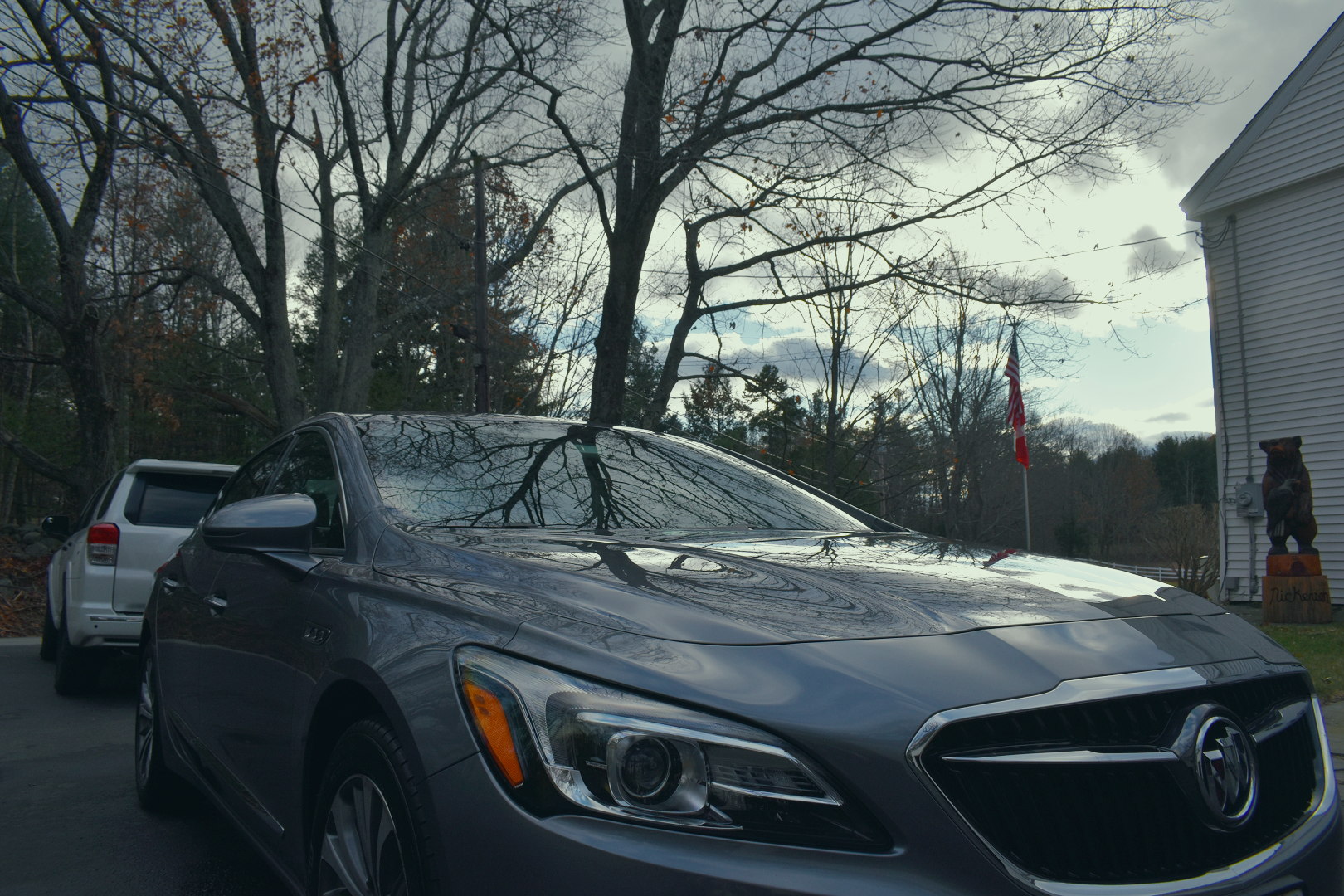A gray car parked outside on a paved surface with trees and a white house in the background. An American flag is flying on a pole next to the house, and a bear statue is positioned near the house wall.