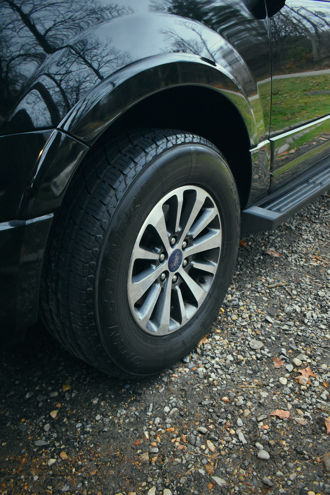 Close-up of a black Ford vehicle's front wheel showing a silver alloy rim and a black tire with the brand name "Dueler A/T" visible. The vehicle is parked on a gravel surface with some fallen leaves, and the background reflects trees and sky on the c