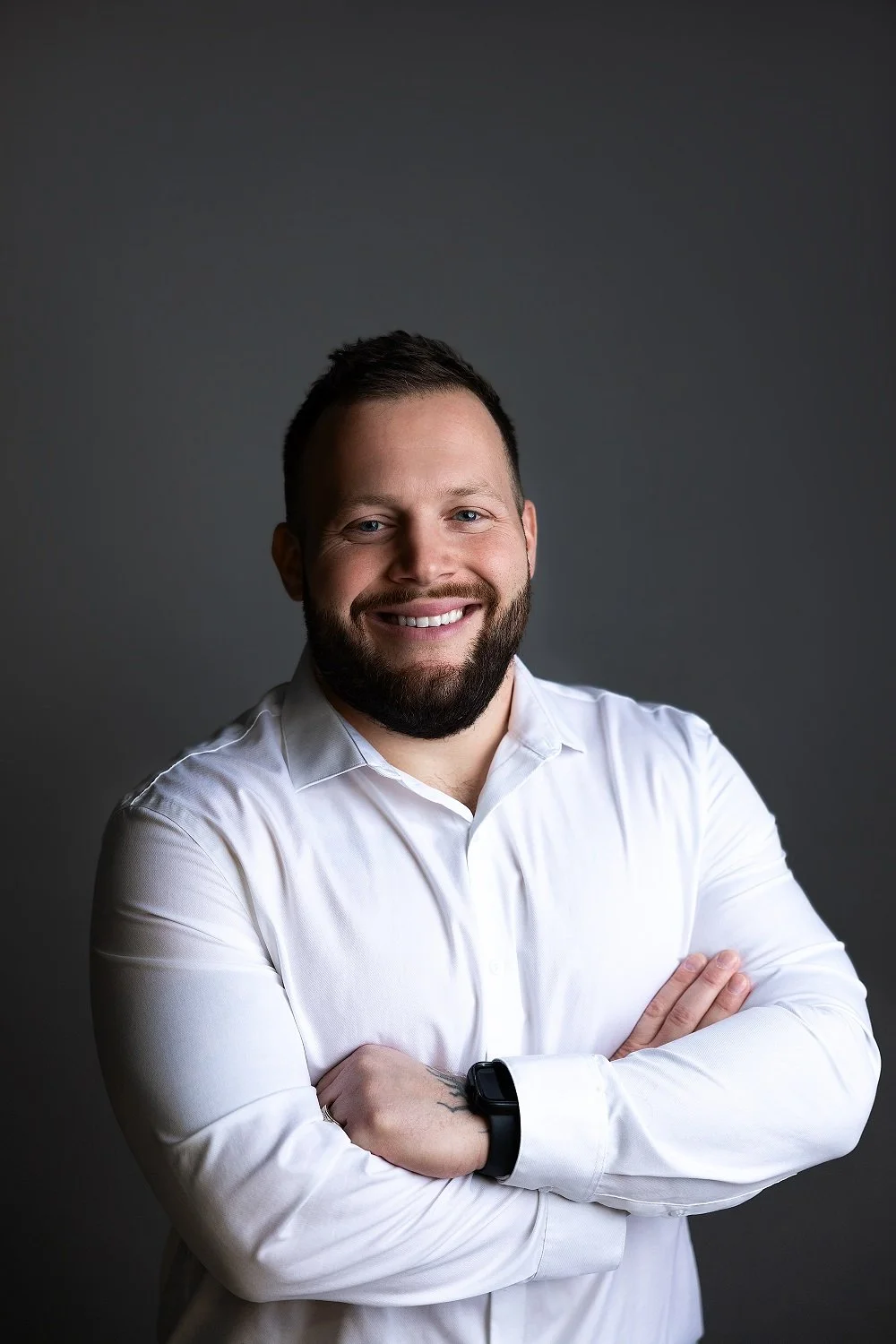 A young man with a beard and short hair smiling with arms crossed, wearing a white dress shirt and a smartwatch, against a gray background.