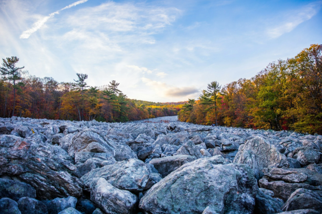 Rocky riverbed with colorful autumn trees on either side and a bright blue sky with light clouds overhead.
