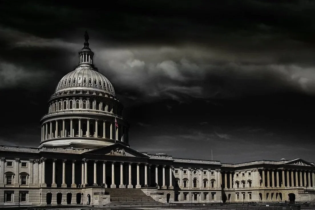 A wide, dark-toned photograph of the United States Capitol, its illuminated dome rising beneath heavy storm clouds that dominate the sky.