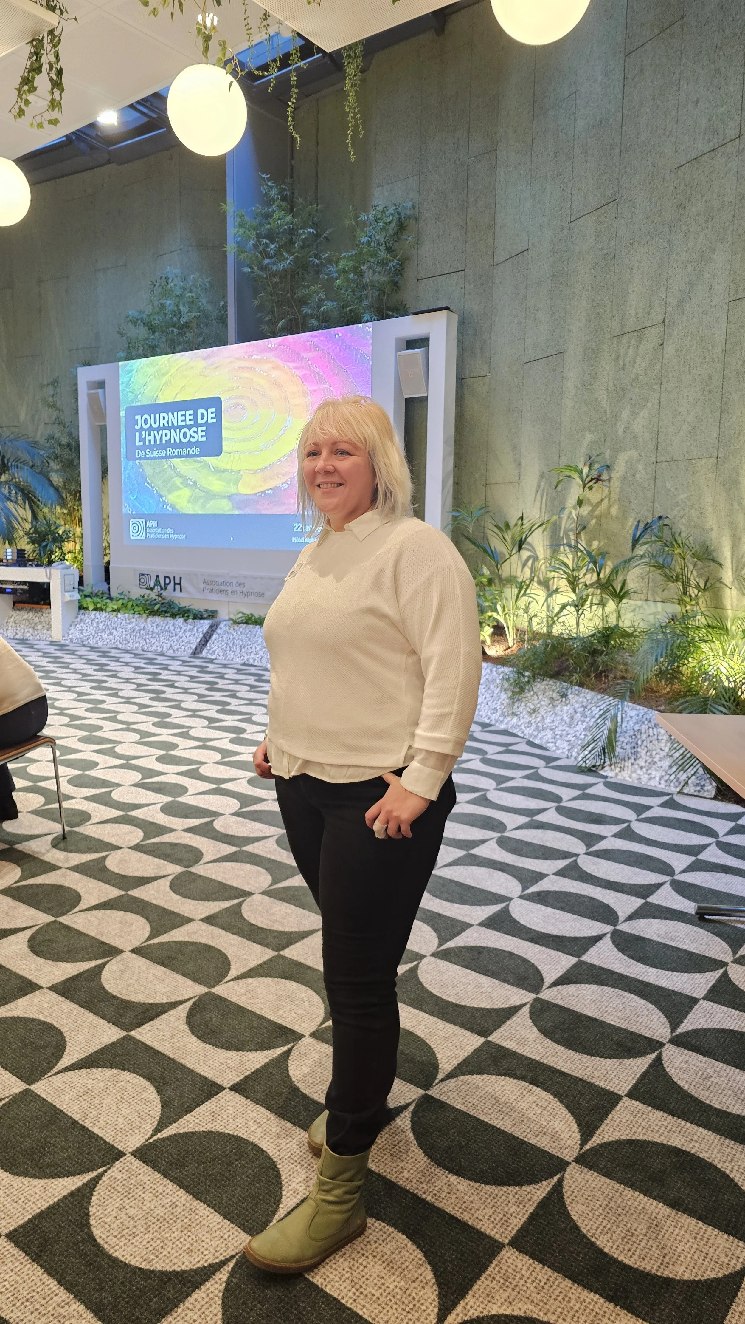 Mia Cosarca debout dans une salle de conférence avec un écran à l'arrière affichant 'Journée de l'hypnose de Suisse Romande'.