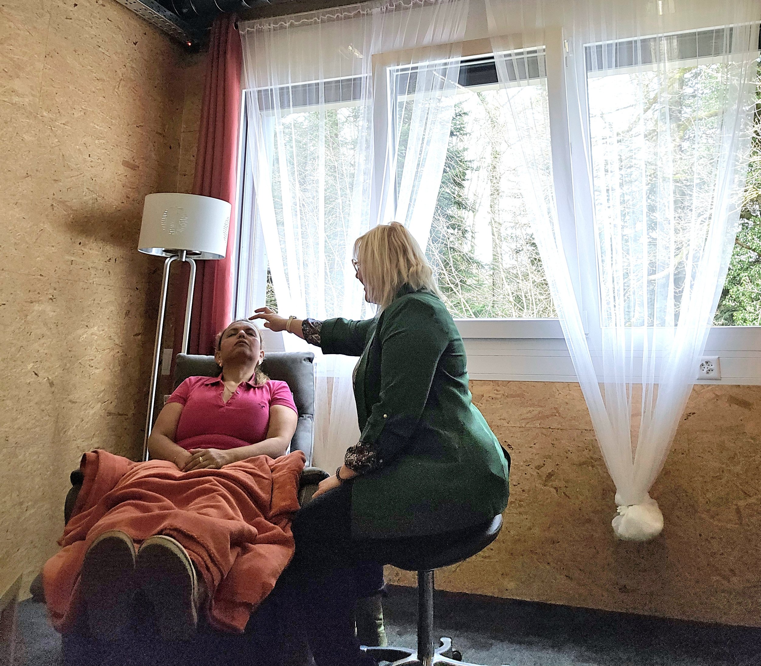 Une femme allongée sur une chaise hypnotisée par Mia Cosarca, dans une pièce avec des fenêtres laissant entrer la lumière.