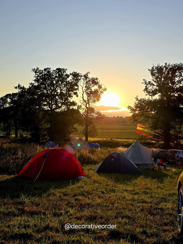Camping tents set up in a grassy field during sunset, with trees in the background and a flag on one of the tents.