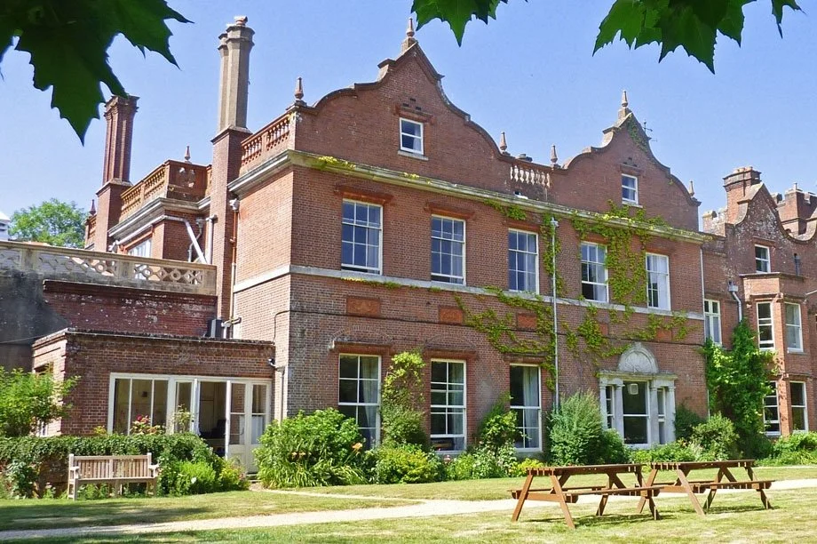 Historic brick mansion with ivy-covered walls, large windows, surrounded by landscaped garden with benches and trees, under a clear blue sky.