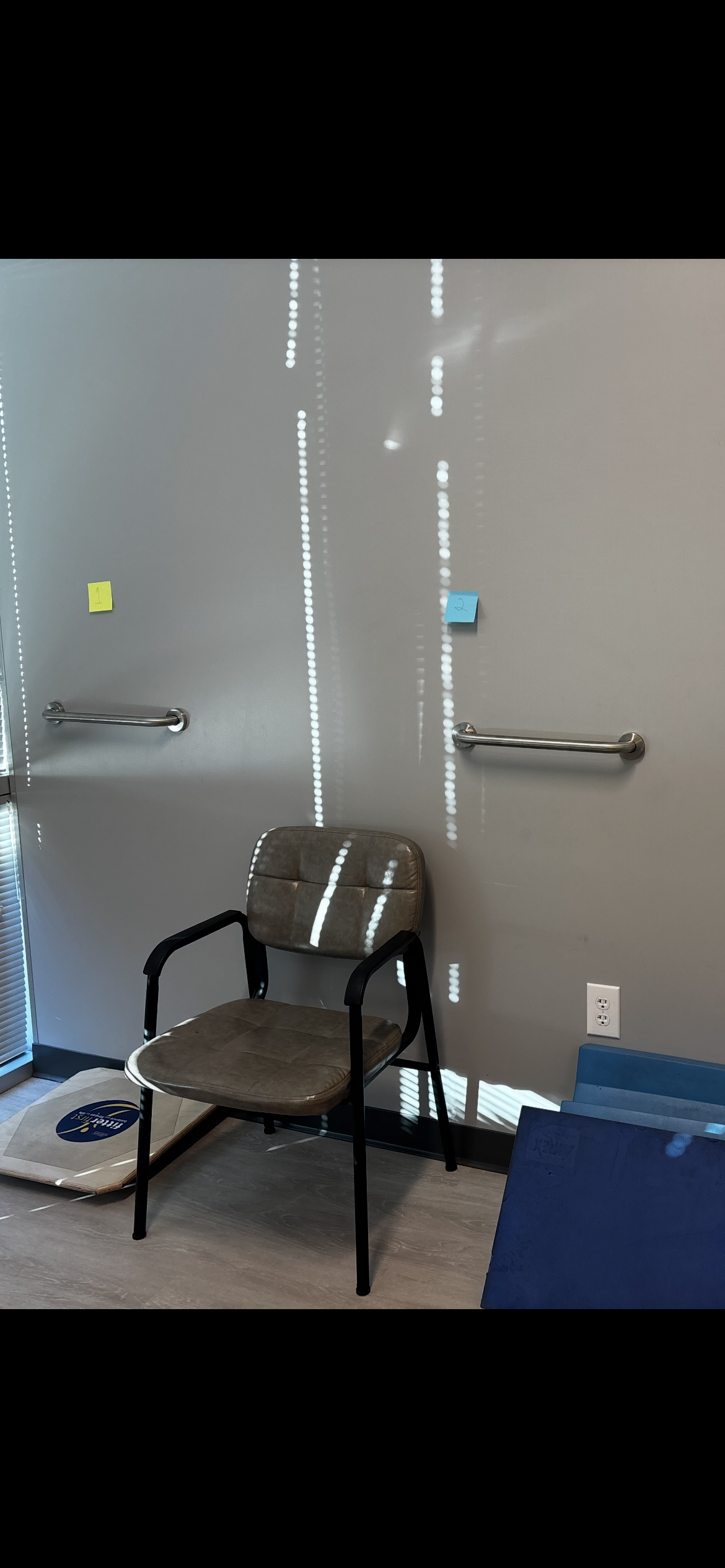 Empty chair in front of a plain wall with two parallel grab bars, one on each side, and small colored sticky notes numbered 1 and 2 on the wall. Natural light creates dotted light patterns on the wall.