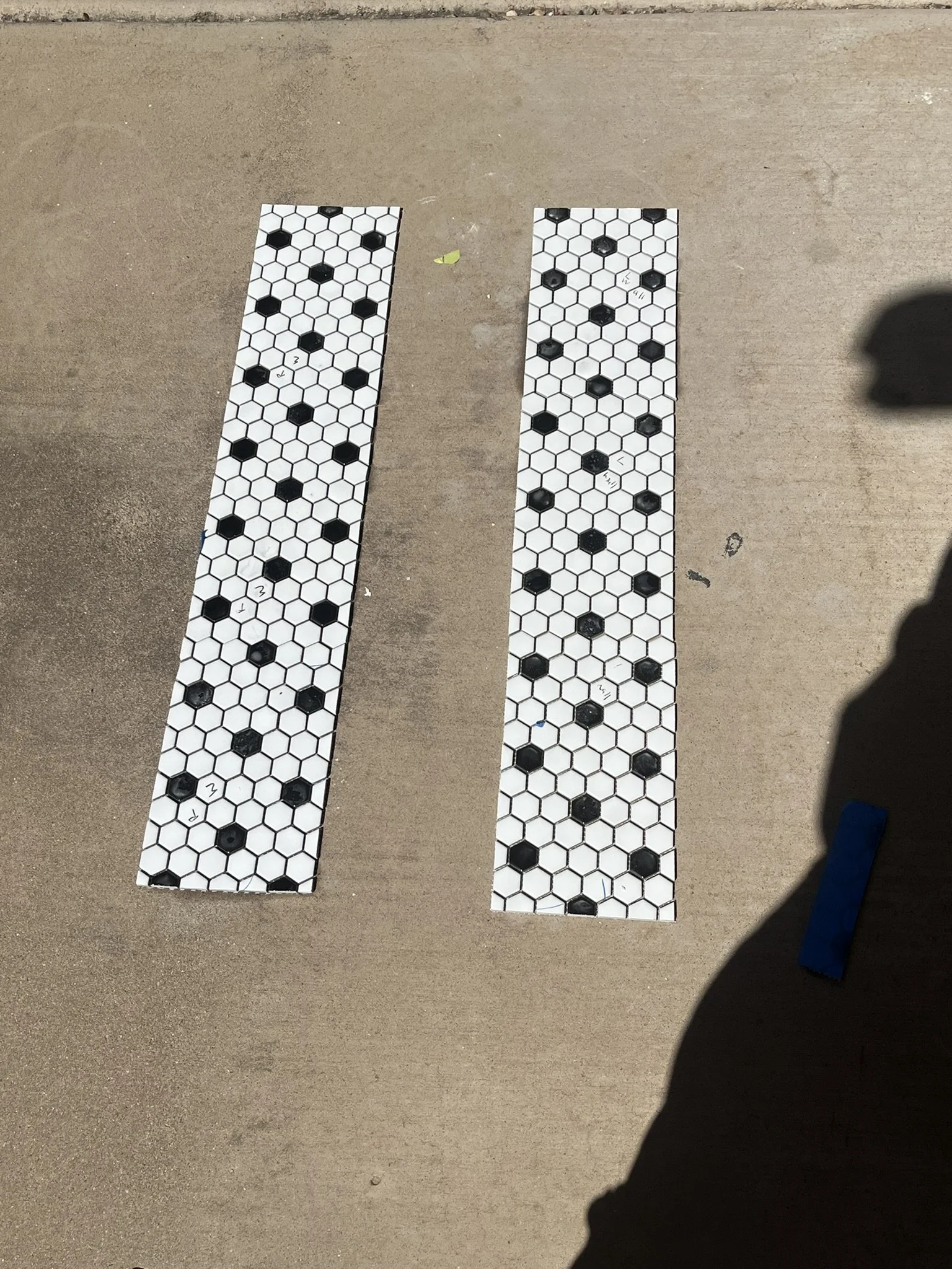 Two narrow strips of white tiles with black hexagon patterns on a concrete surface, with some tiles marked with letters and drawings, and shadows cast on the right side.