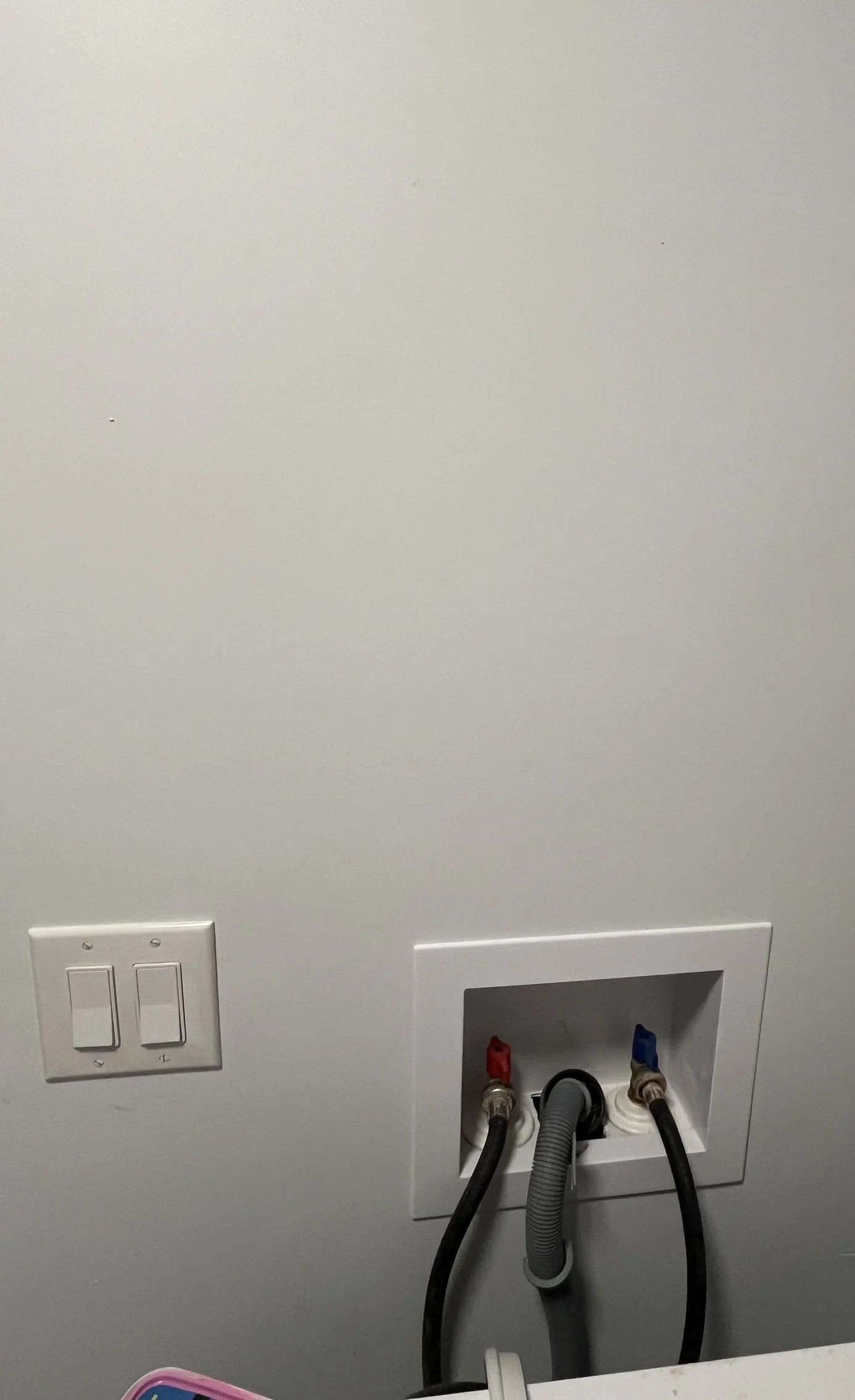 Wall with electrical outlets and water supply connections, one with black hoses attached, in a laundry area.
