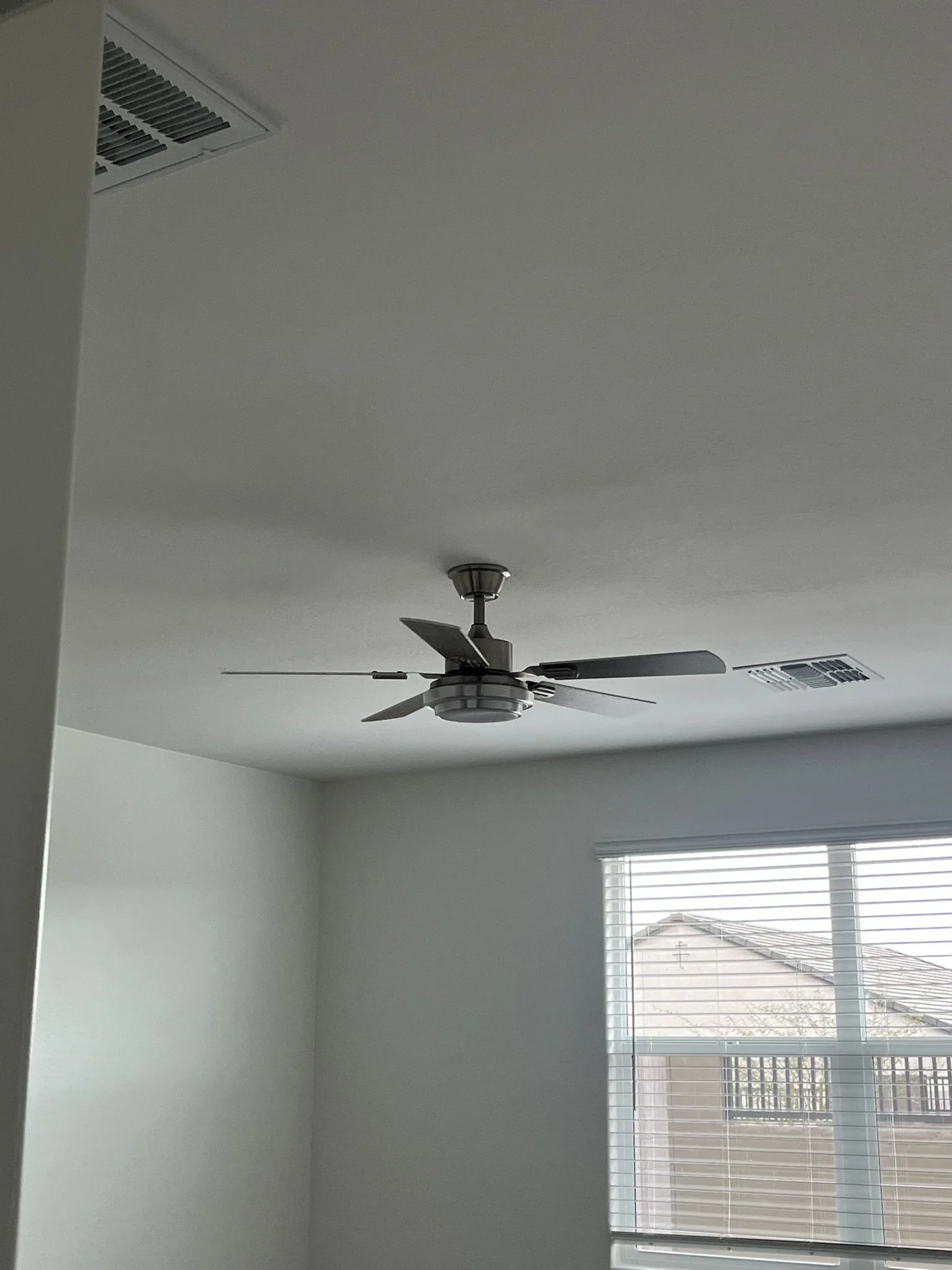 Empty room with white walls, a ceiling fan, a large window with blinds, and two air vents on the ceiling.