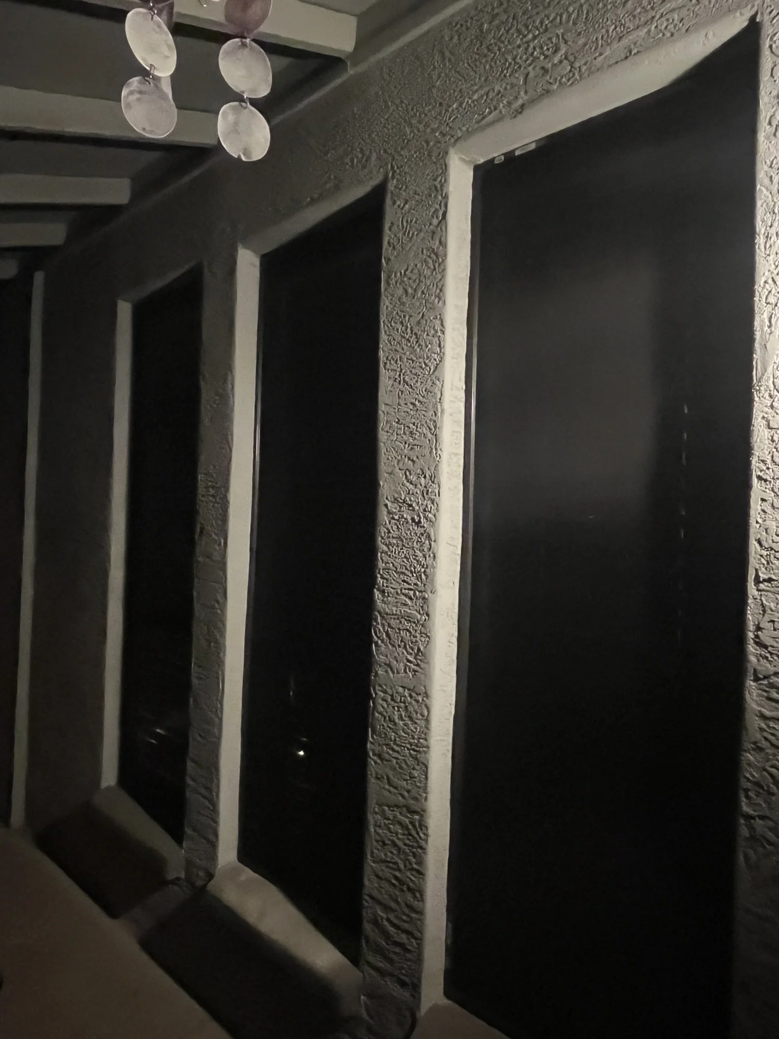 Dimly lit interior showing three large square window openings in a textured wall, with a string of white spherical decorative objects hanging from the ceiling.