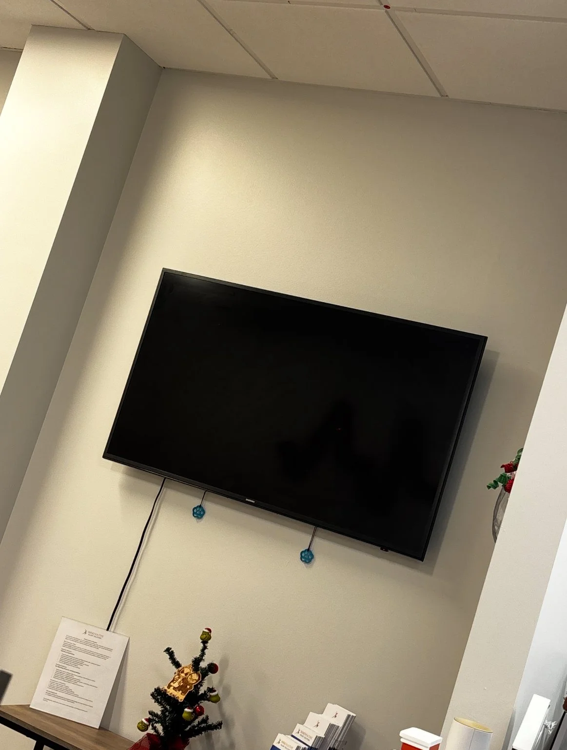 A mounted flat-screen television on a white wall with two blue decorative hanging charms below it, a small Christmas tree with ornaments, and some informational papers and small items on a wooden table.