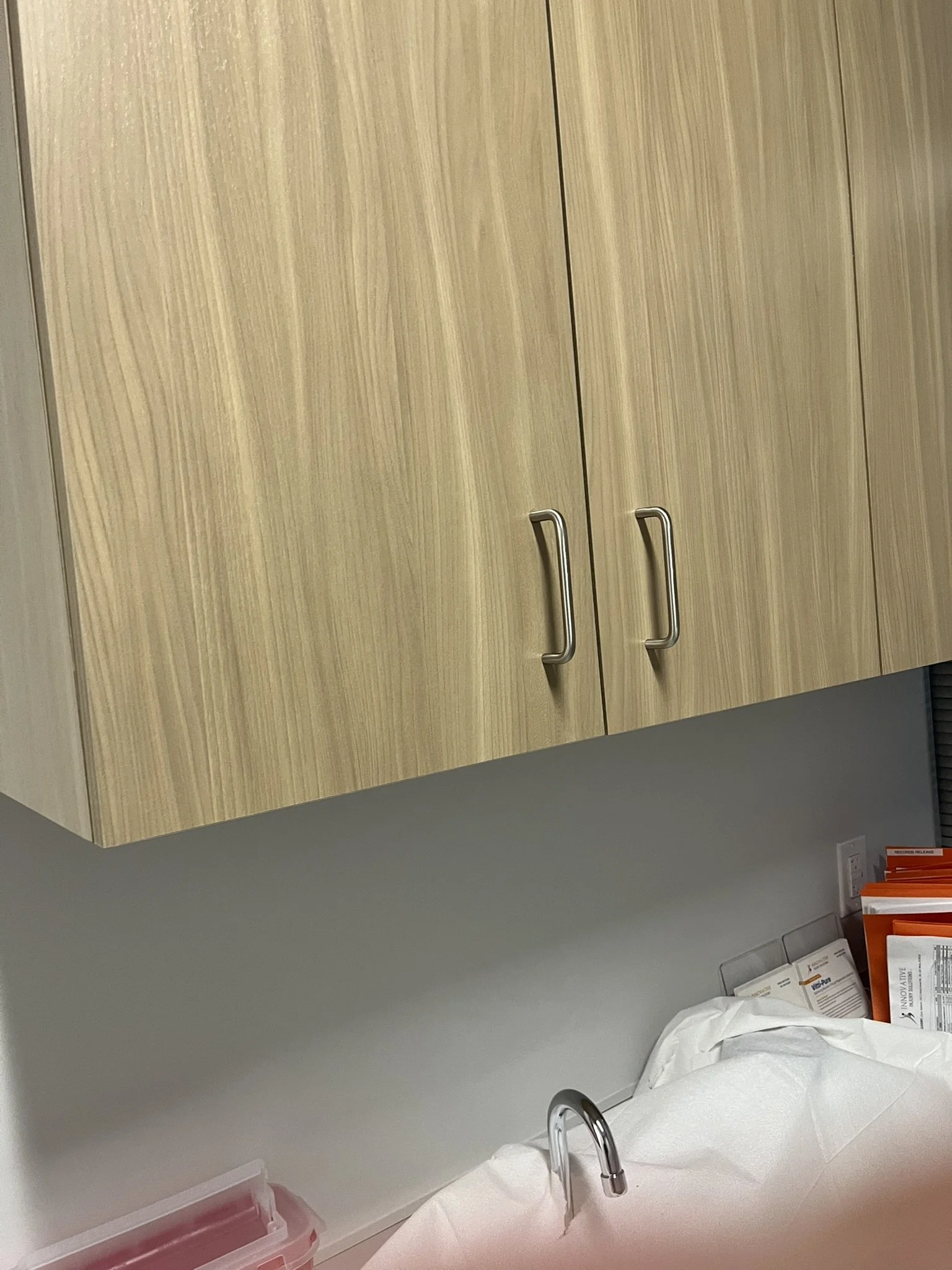 Wooden storage cabinet with silver handles above a white countertop with a sink, and some orange and white documents or folders on the right side.