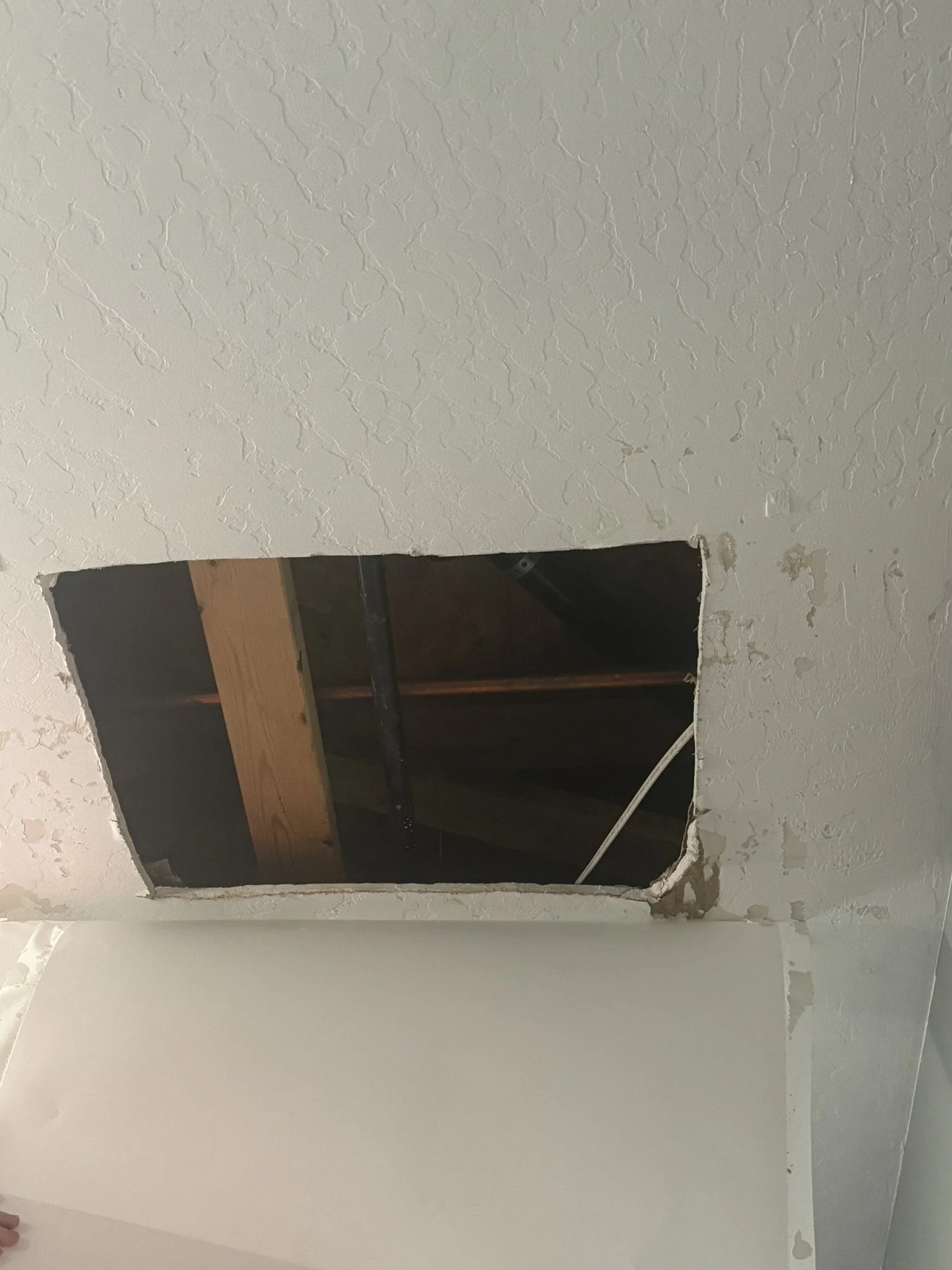 Ceiling with a square hole exposing wooden beams and pipes.