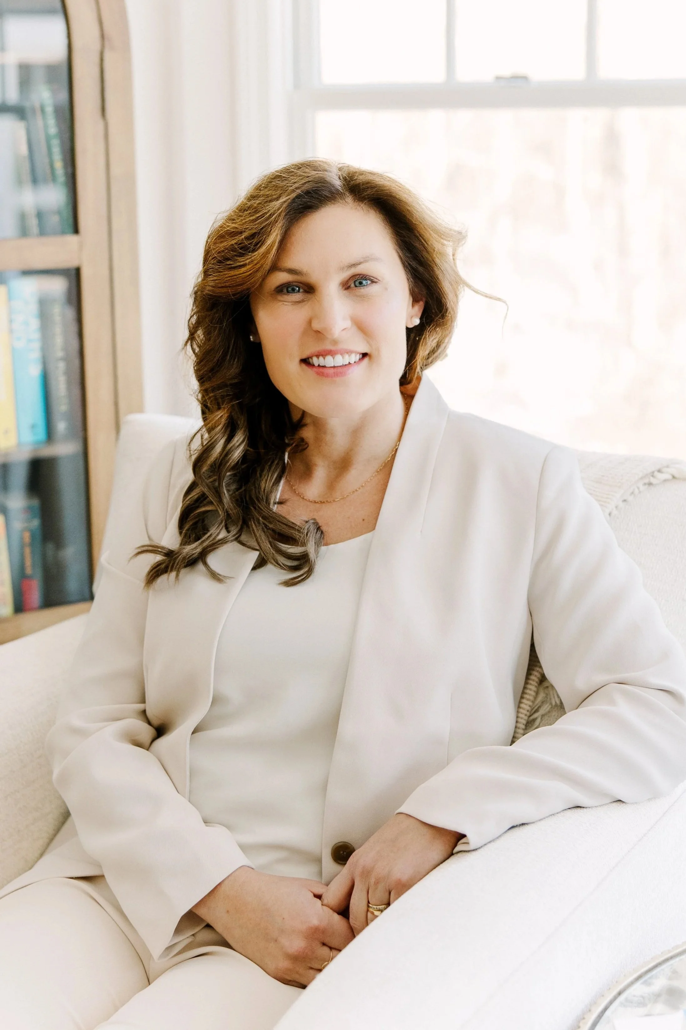 A woman with wavy brown hair, smiling, sitting on a cream-colored sofa, wearing a white blazer and matching top, with a bookshelf and window in the background.