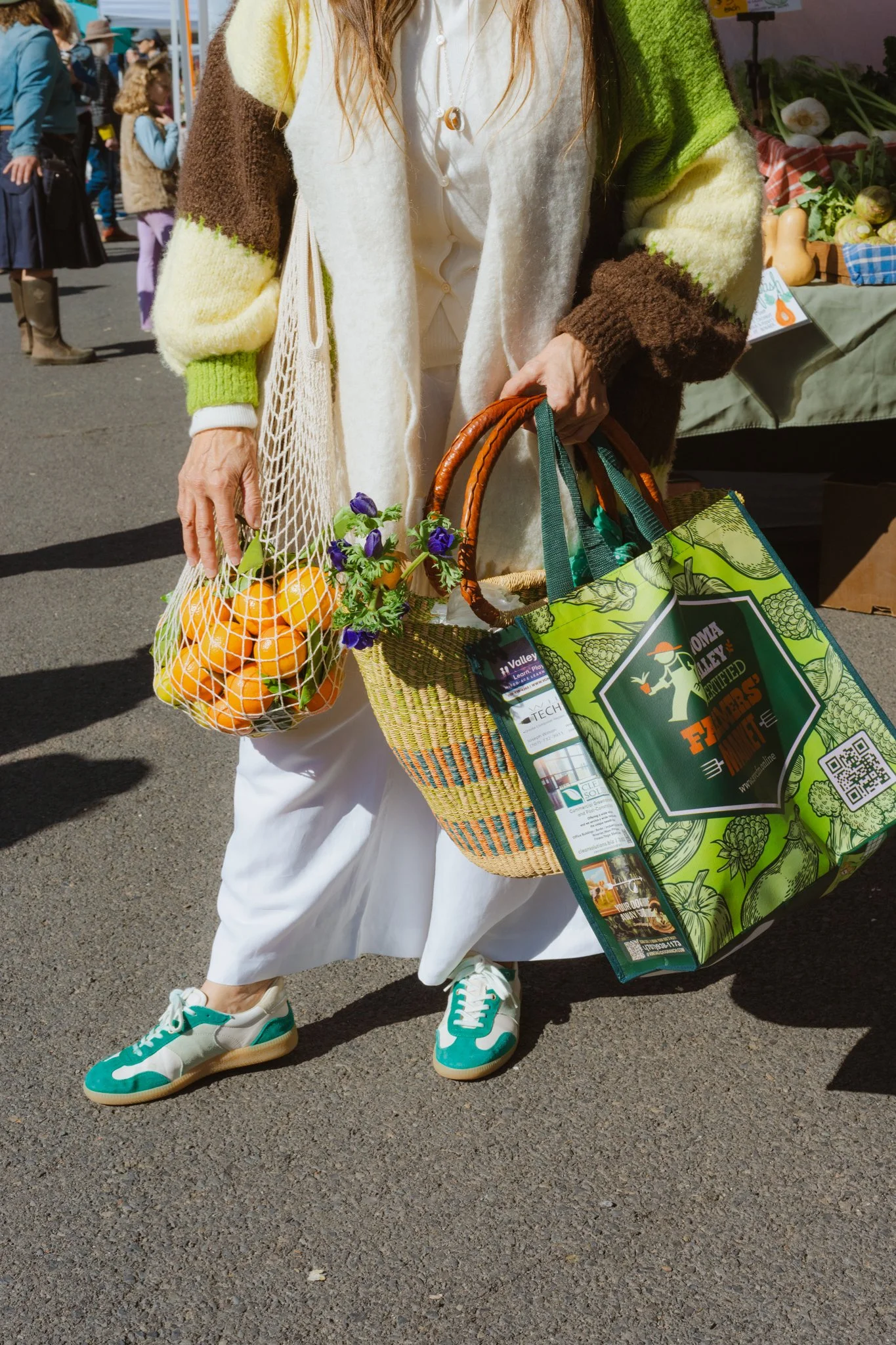 Person holding a farmers market bag filled with oranges, a small bouquet of purple flowers, and shopping bags at an outdoor market, with other people and market stalls in the background.