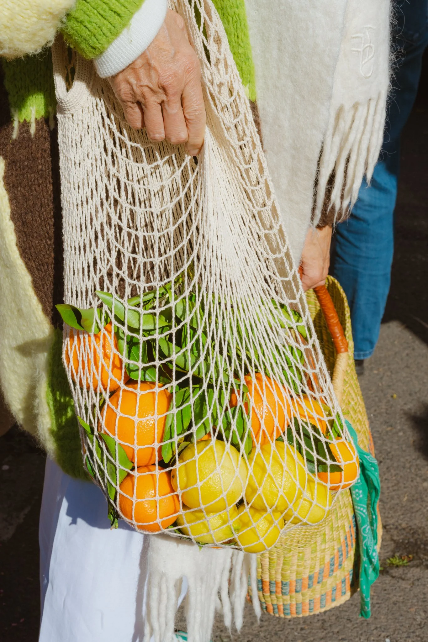 A person holding a farmers market bag filled with oranges and lemons, wearing a colorful scarf.