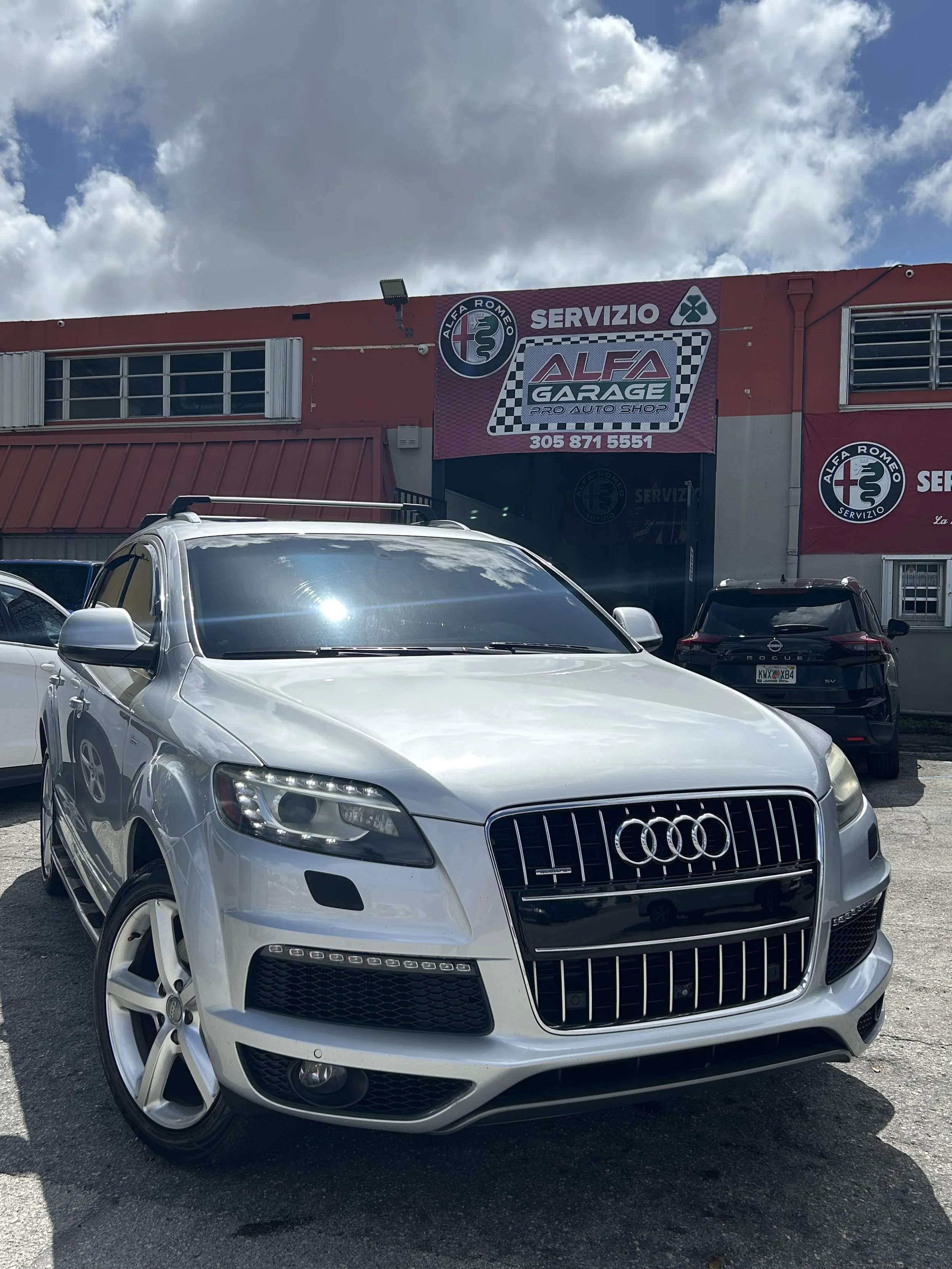 Front view of a silver Audi sedan parked in a parking lot with other vehicles, against a background of a building with a sign for Alfa Garage auto shop.
