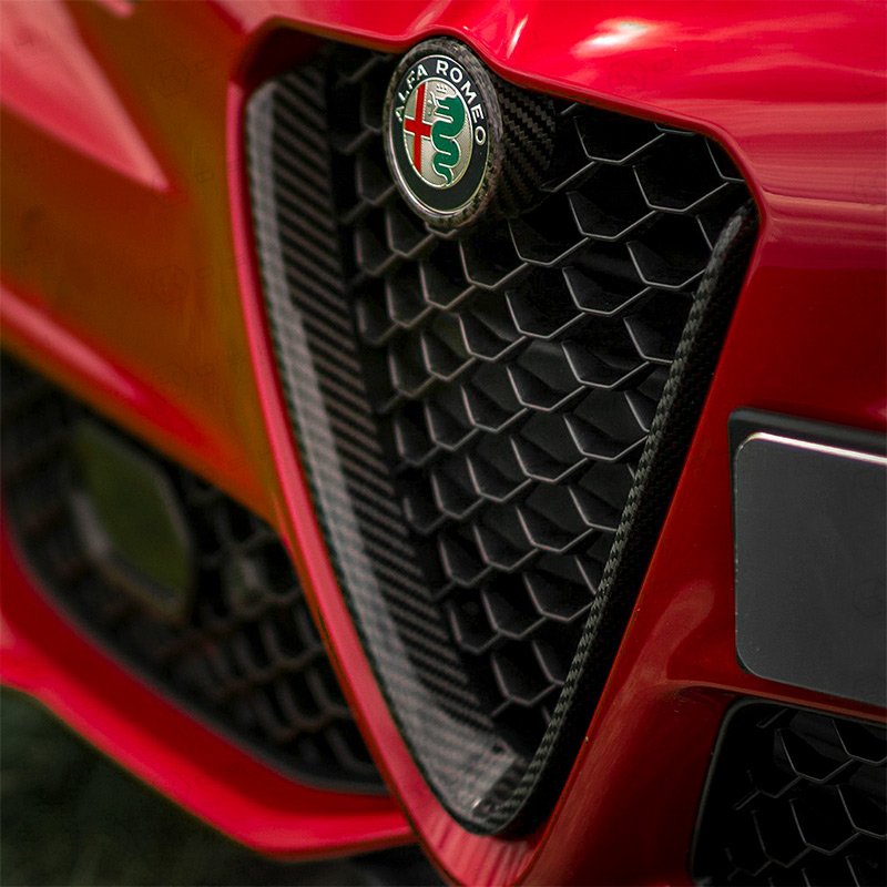 Close-up of the front grille of a red Alfa Romeo car, featuring the Alfa Romeo badge and hexagonal honeycomb design.