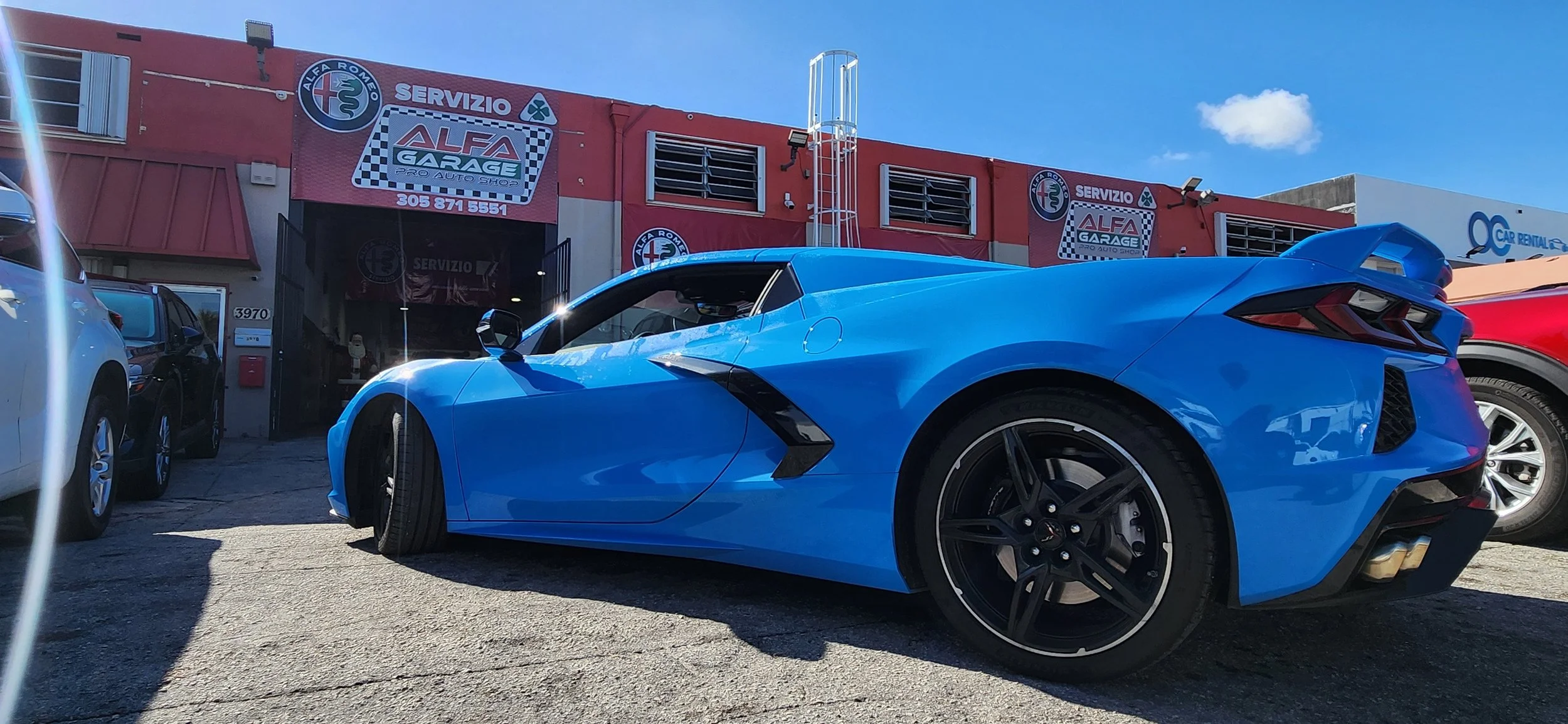 A bright blue sports car parked outside an auto repair shop with a red facade, signage for Alfa Garage, and several other vehicles nearby.