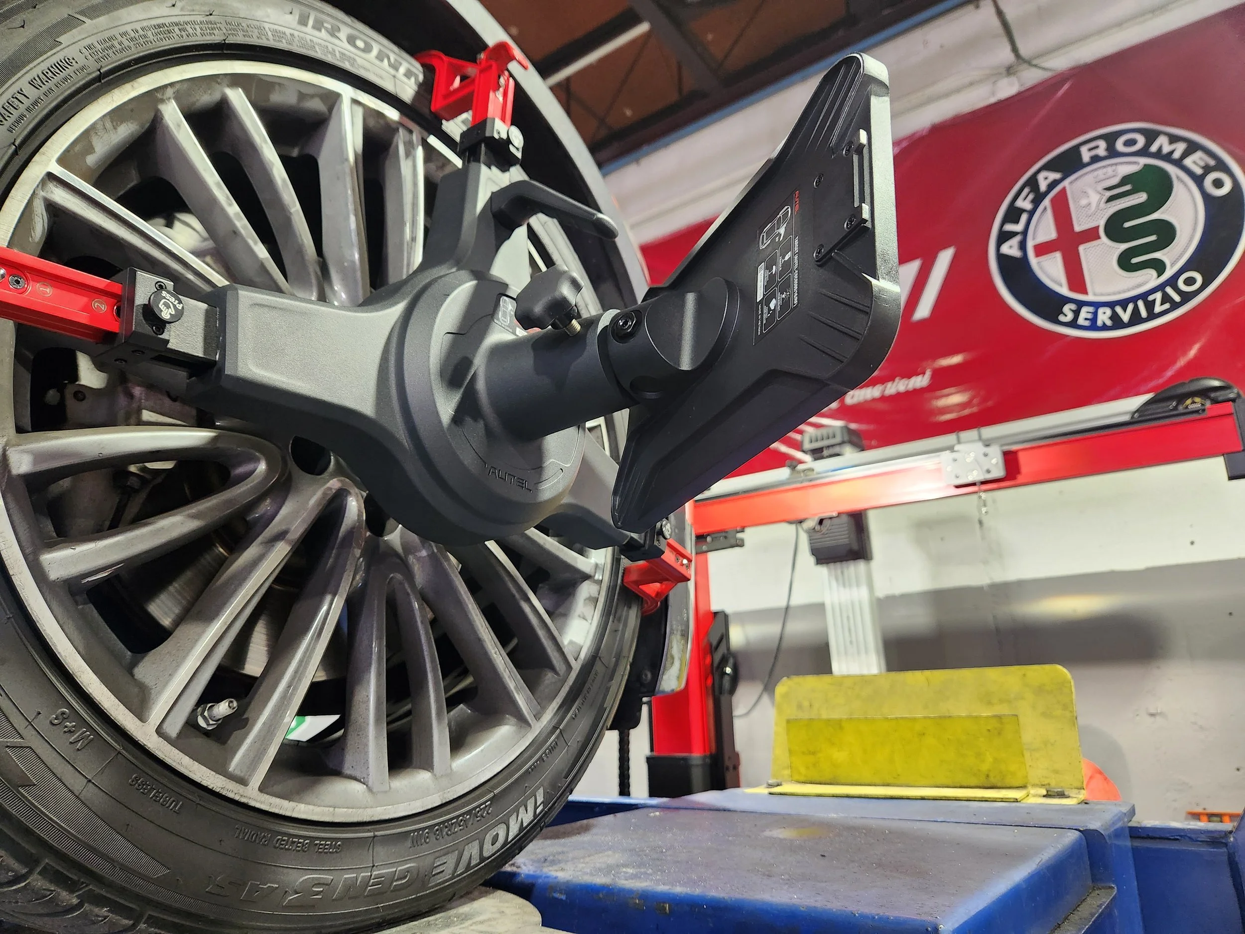 Close-up of a wheel and tire mounted on a car in a garage, with a tire pressure monitoring system sensor attached to the wheel. In the background, there is a red tool chest with an Alfa Romeo sign on the wall.