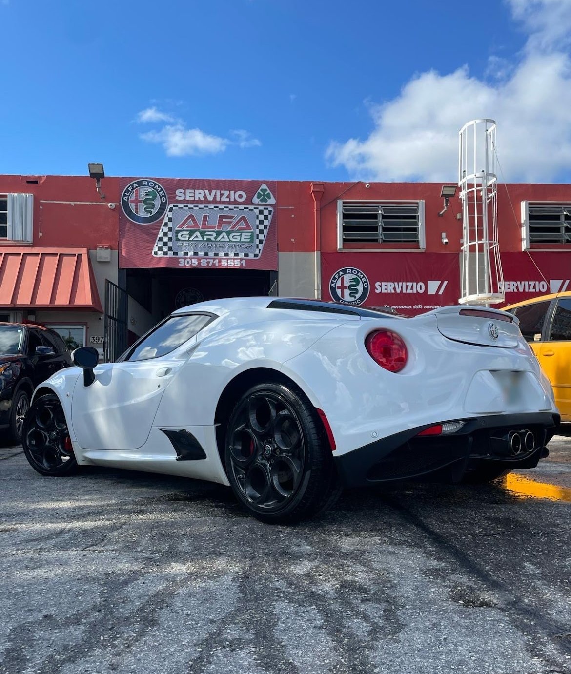 White sports car parked outside Alfa Romeo service garage with a red building, blue sky, and other parked vehicles.